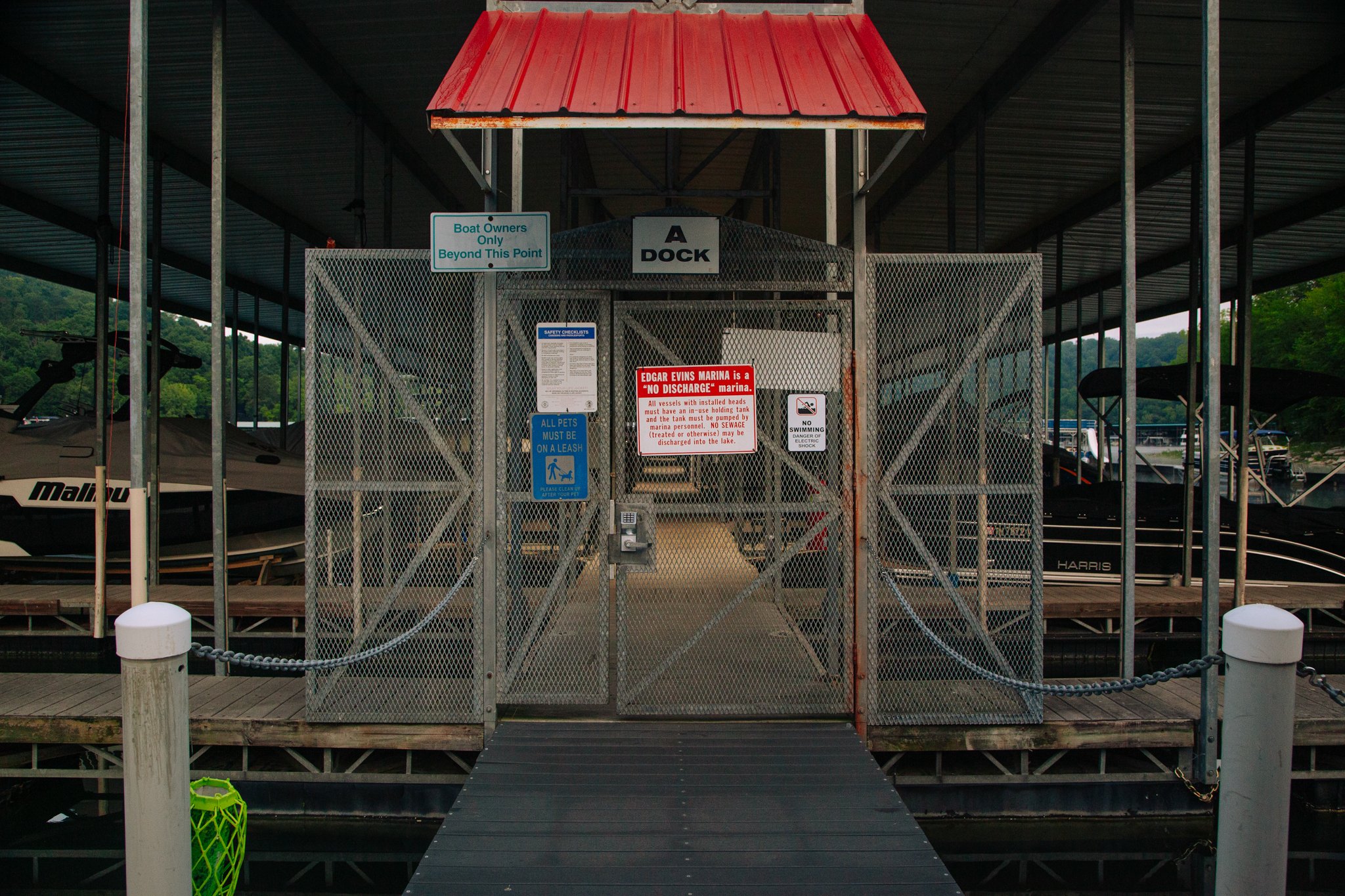 A dock with a gated entrance and signs, including one that says 'Boat Owners Only Beyond This Point' and another indicating 'No Discharge.' There is a boat in the background and a ramp leading to the water.