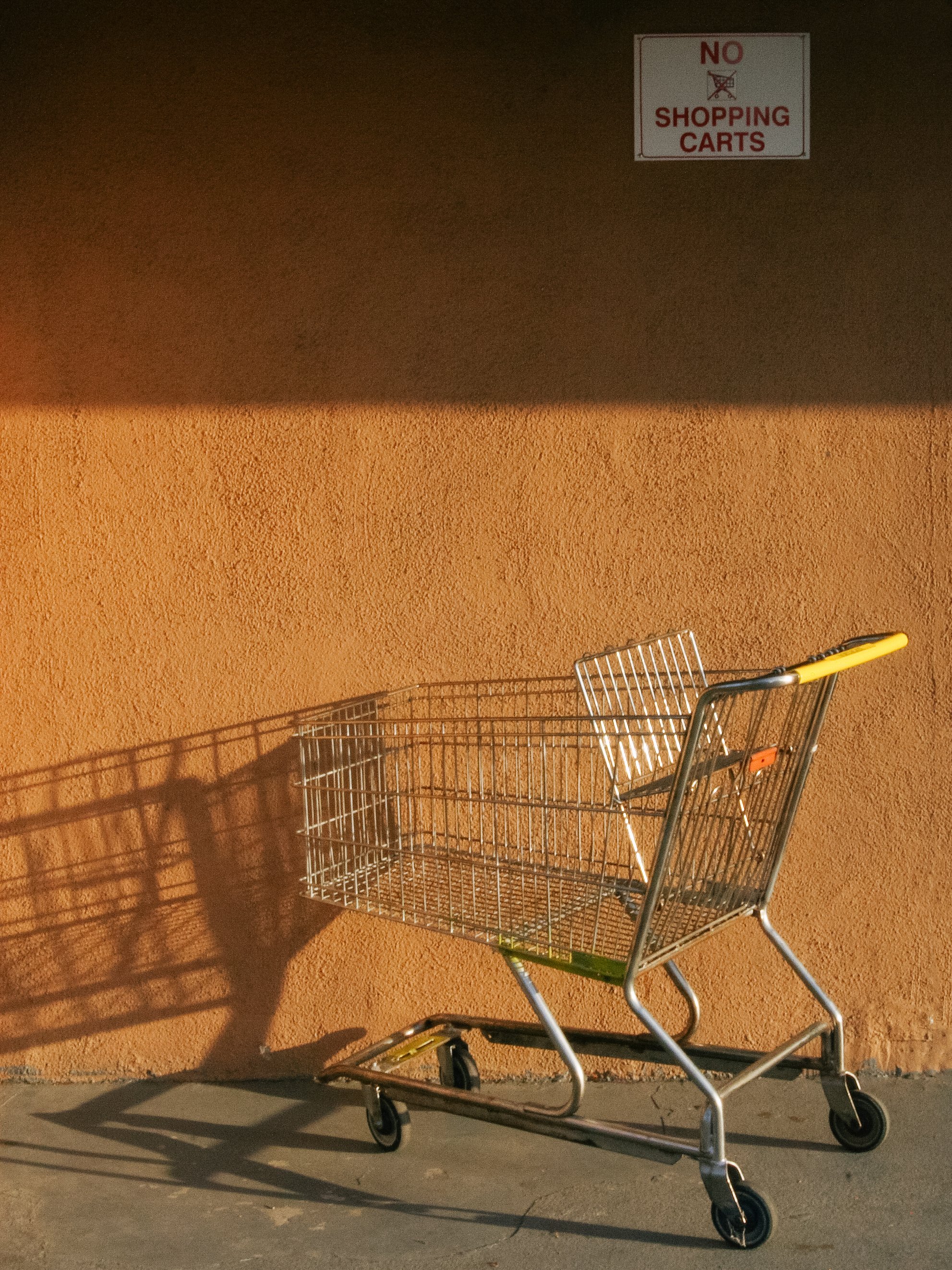 Empty shopping cart with yellow handlebar casting a shadow on an orange wall, with a sign above that reads 'No Shopping Carts'.