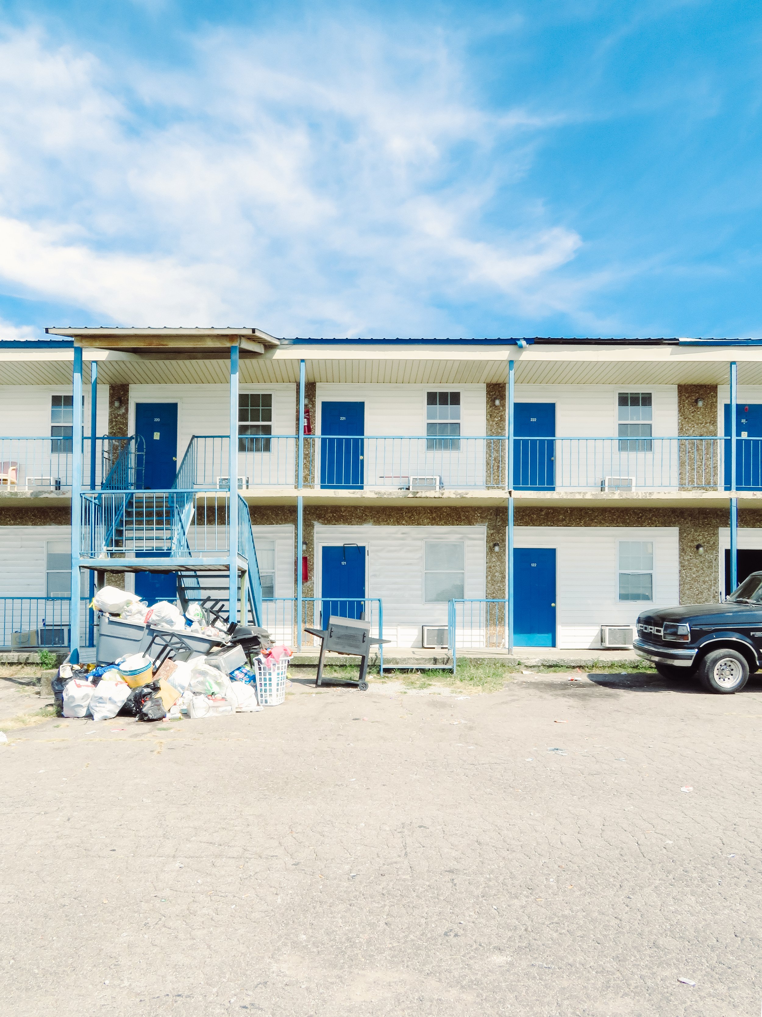 Exterior view of a two-story apartment building with blue doors, windows, and stair railings, under a partly cloudy sky, with a pile of trash and debris in the foreground and a parked black truck on the right side.