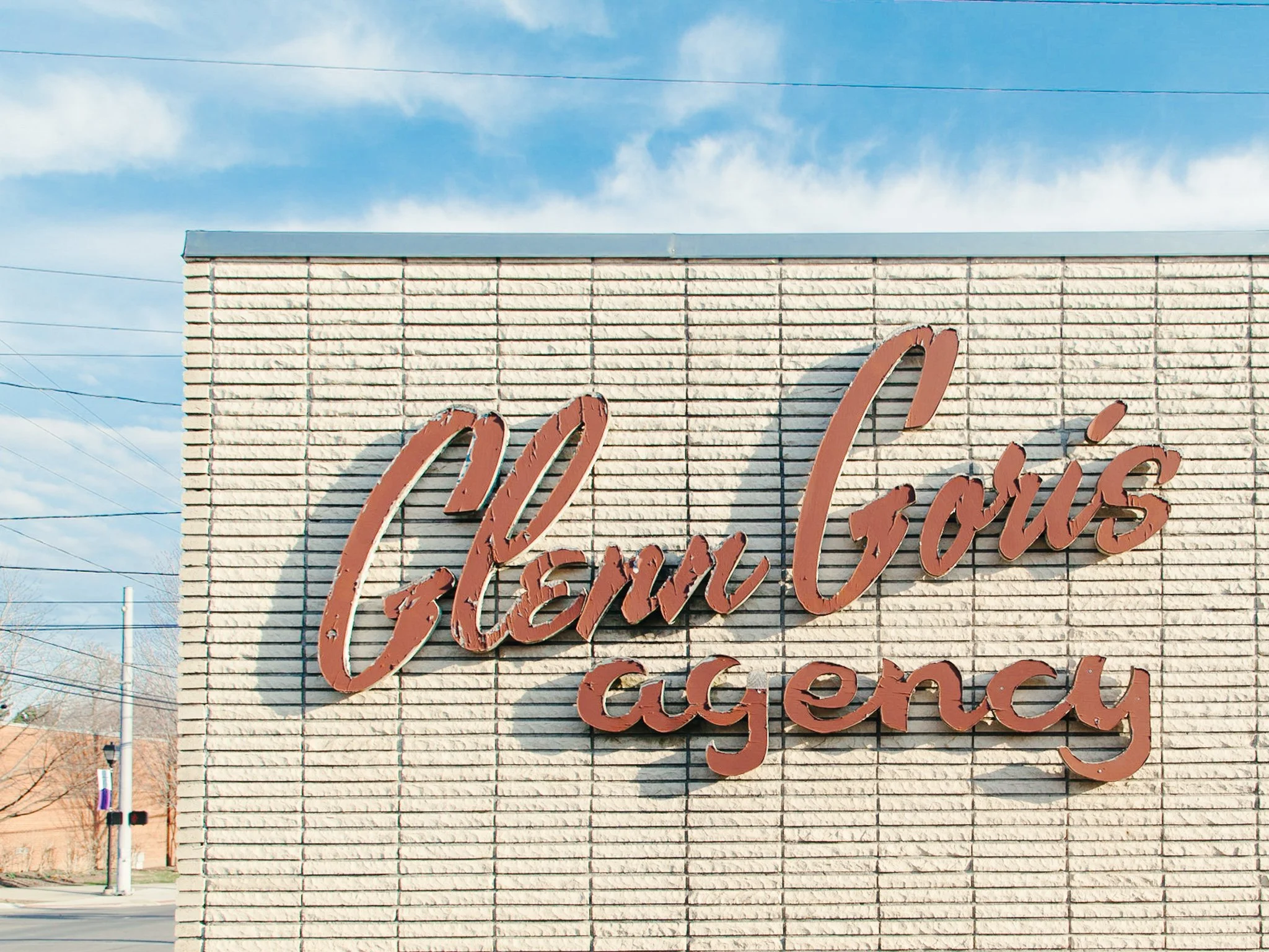 Close-up of a beige brick building with a partial view of a neon sign reading 'Gourmet' in cursive lettering, against a blue sky with clouds and power lines.