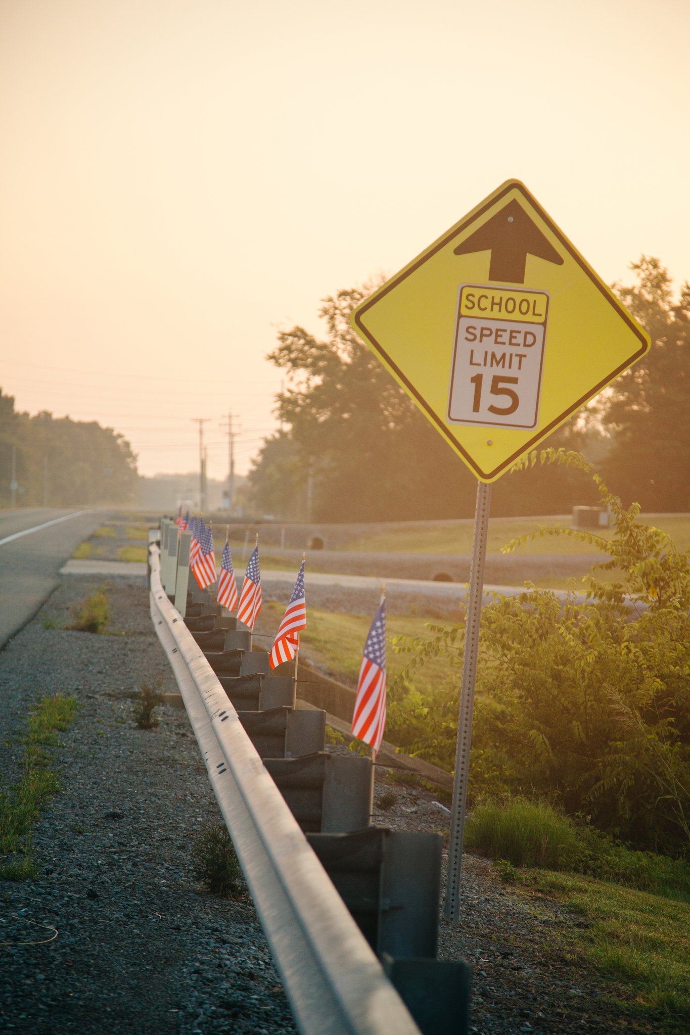 Roadside sign indicating a school zone with a speed limit of 15 miles per hour, and an arrow pointing straight ahead, at sunset. American flags are displayed along the roadside.