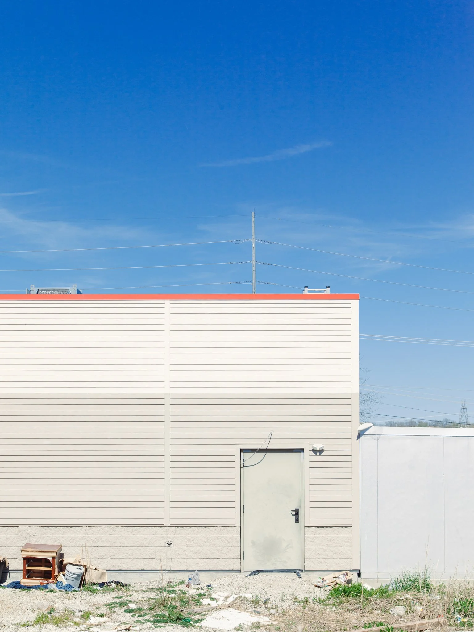 Exterior of a modern building with beige siding, a white door, and minimal landscaping, under a clear blue sky with visible power lines.