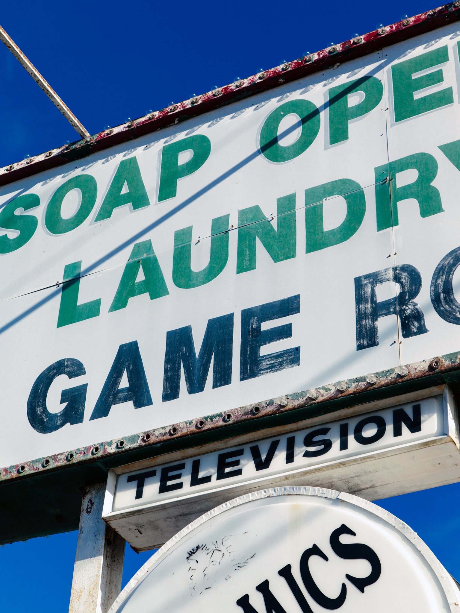 Close-up of an old, weathered sign with the words 'SOAP OPEN LAUNDER' and part of another sign below with the word 'TELEVISION'. The signs are worn with rust and peeling paint, against a clear blue sky.