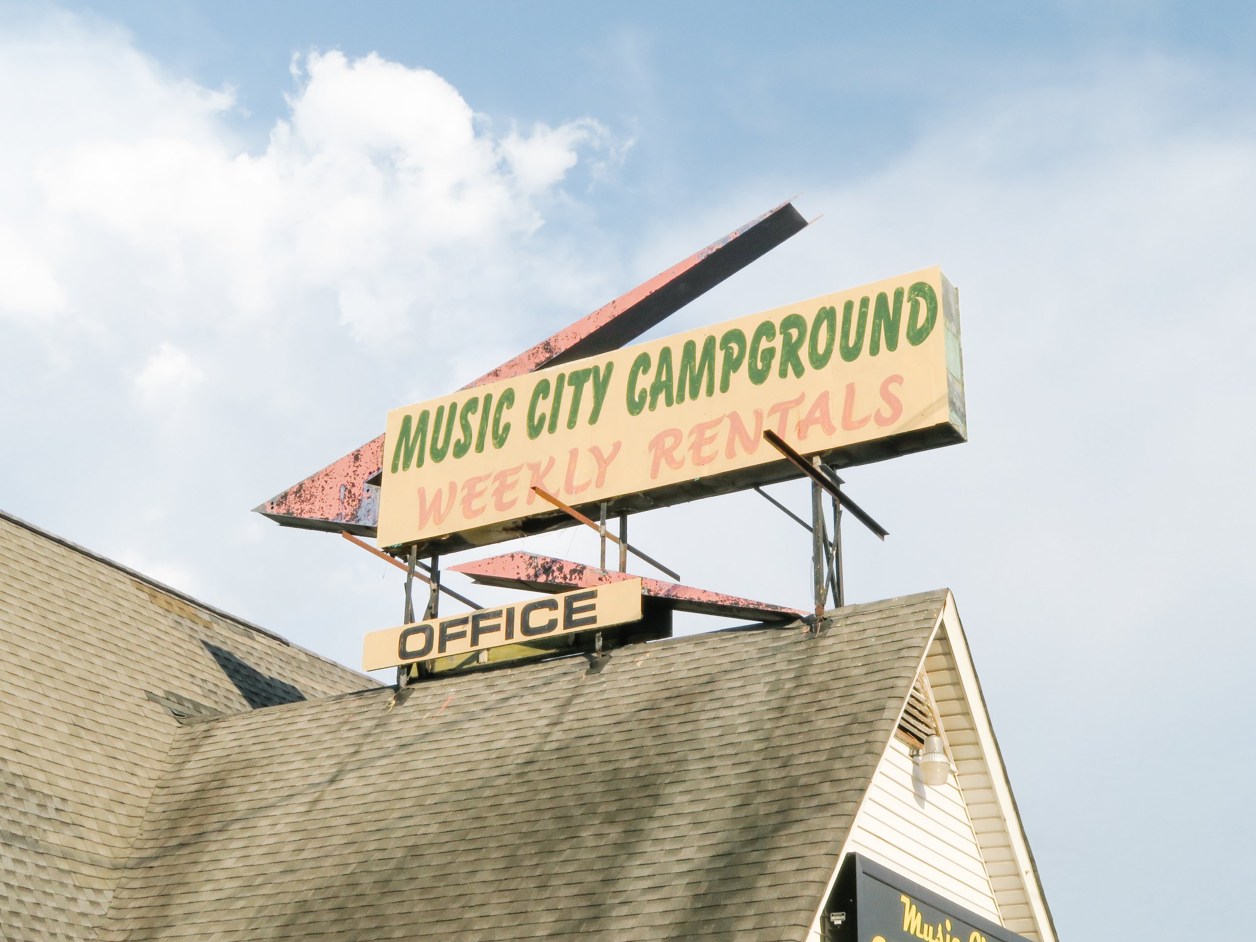 A large sign with green and red text on a yellow background reading "Music City Campground Weekly Rentals" was mounted on metal supports above a building with a shingled roof. The sky in the background is partly cloudy.