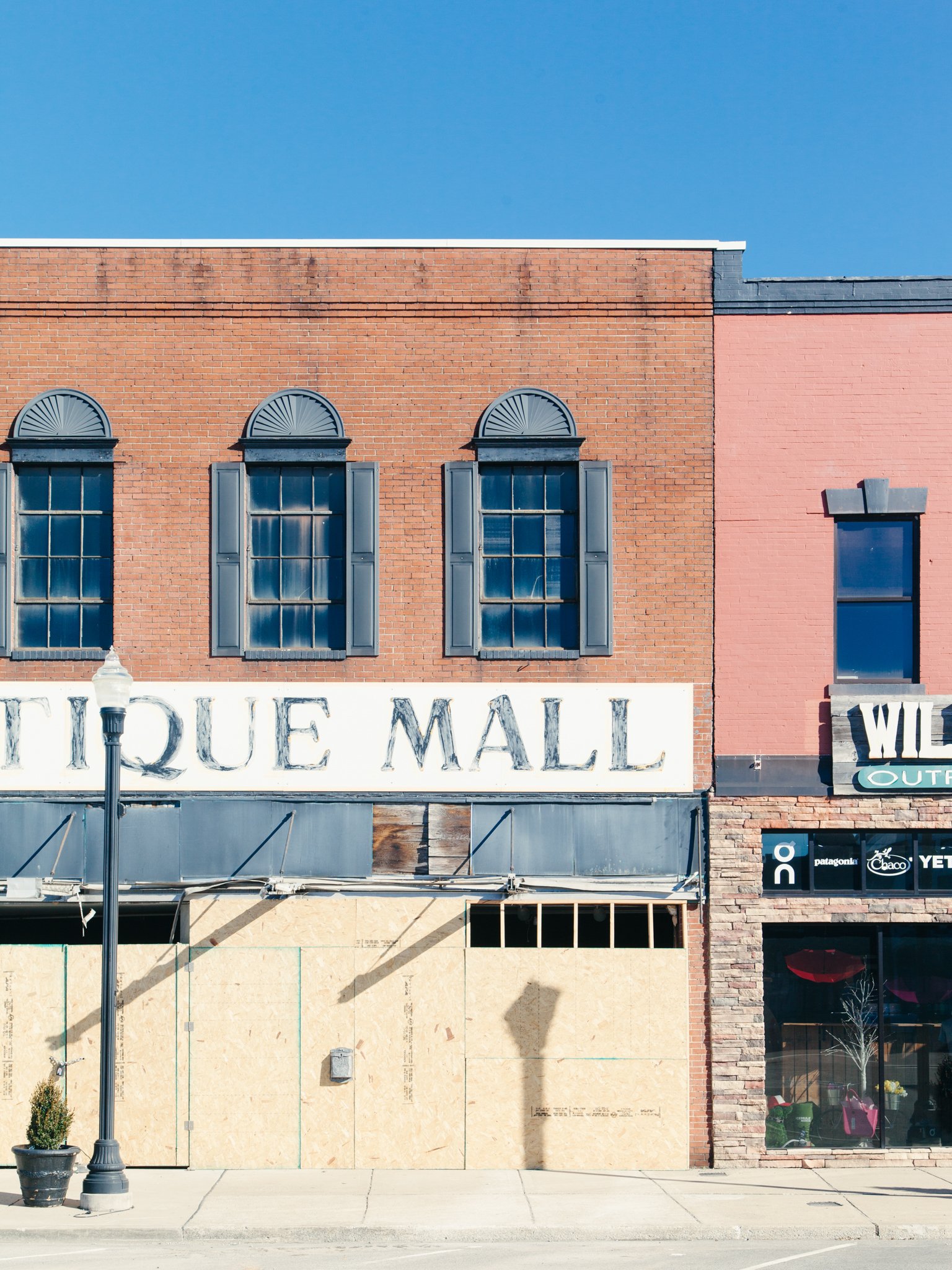 A brick building with three tall windows and blue shutters, partially covered by a boarded-up storefront, under a clear blue sky.