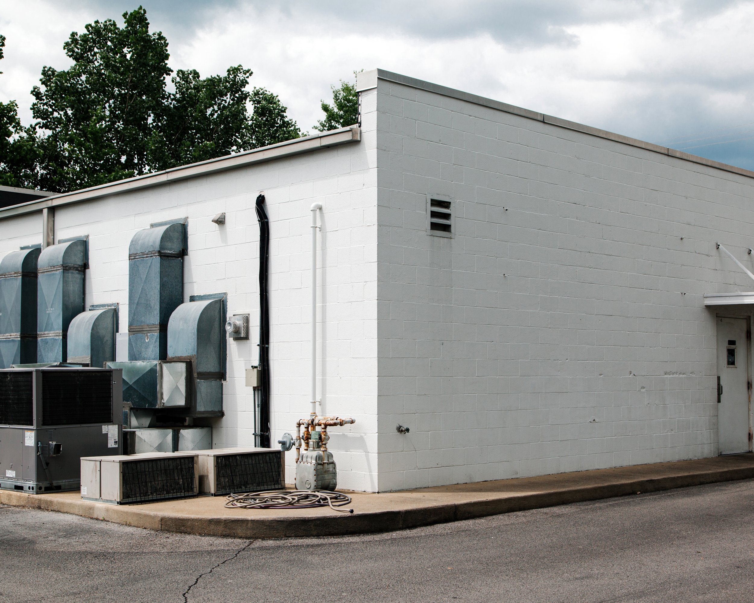 Back of a white commercial building with HVAC equipment and vents, surrounded by a parking lot and trees under a cloudy sky.