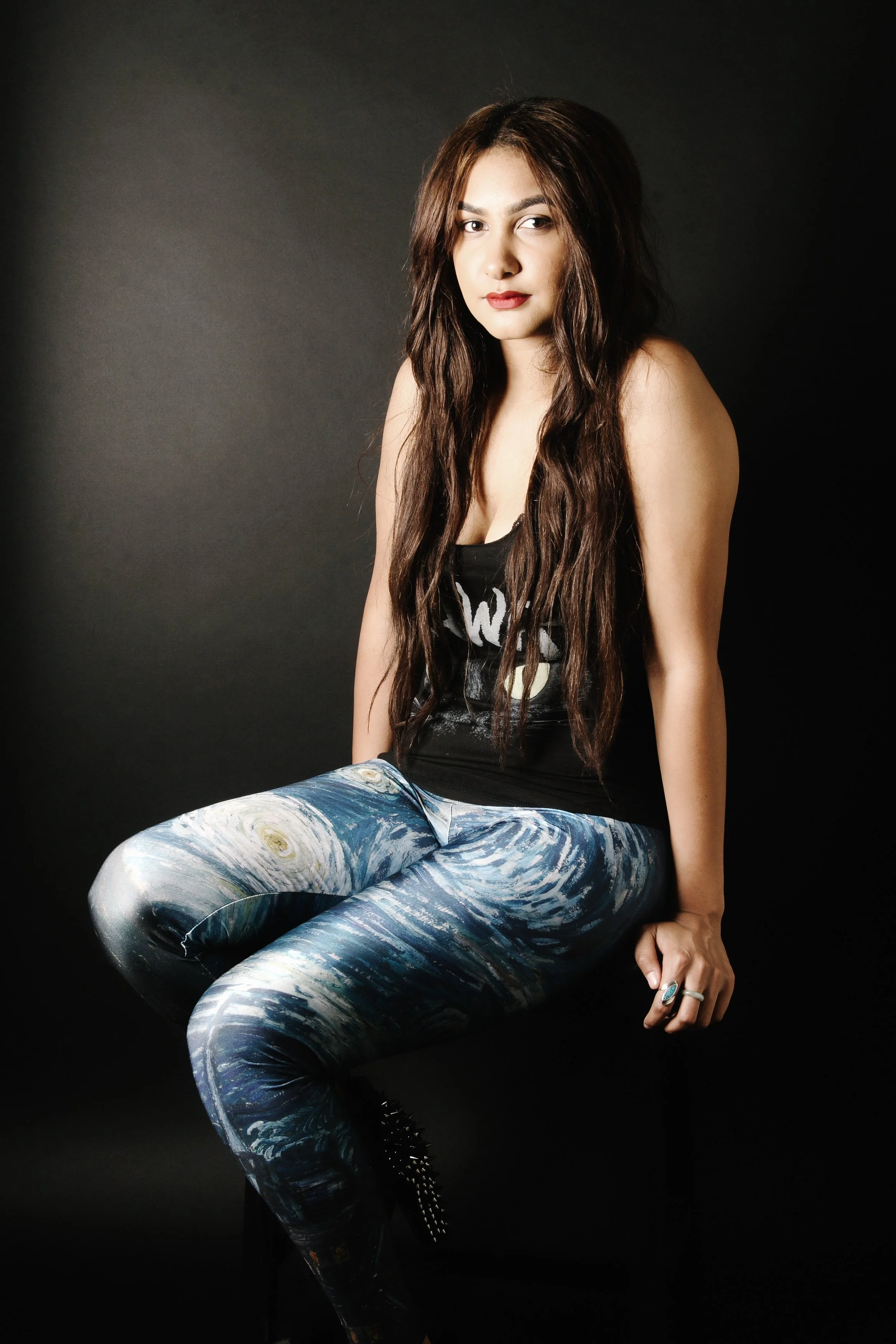Young woman with long wavy brown hair sitting against a black background, wearing a black sleeveless top with white writing and colorful abstract patterned pants.