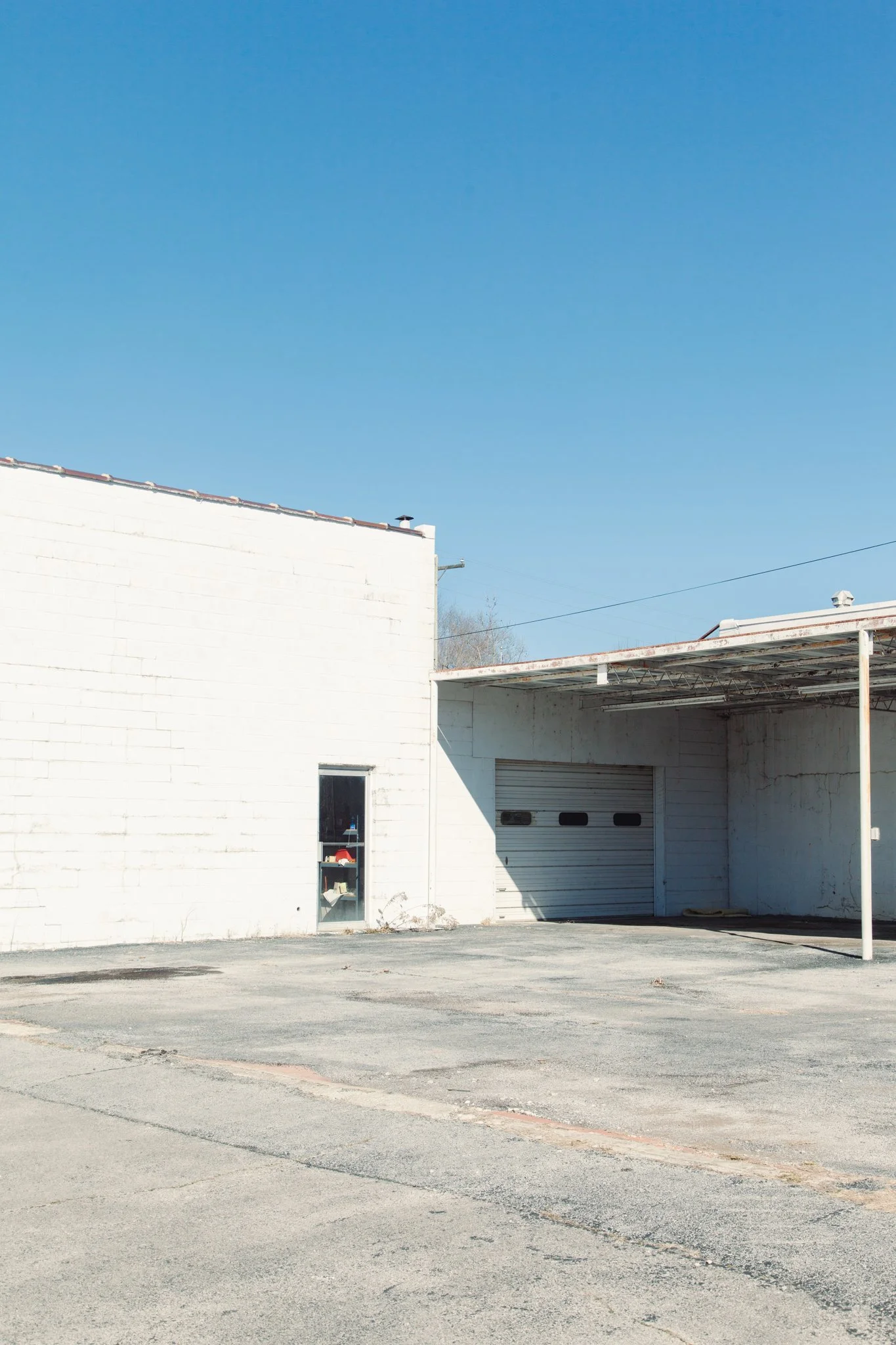 A plain white building with a closed garage door and small window, on a cracked concrete lot with no vehicles, under a clear blue sky.
