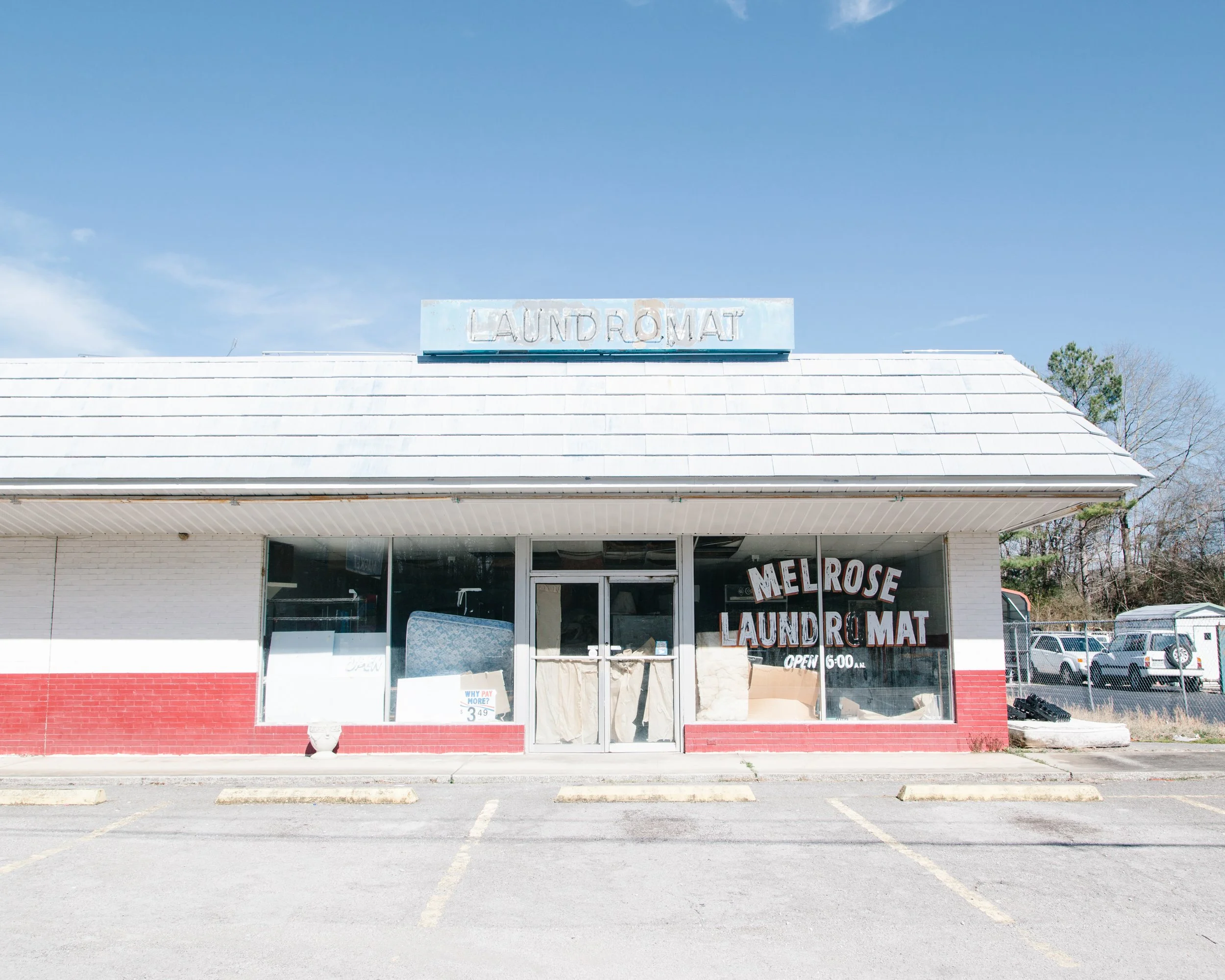 A small laundromat with a sign on the roof reading 'LAUNDROMAT' in faded letters, large glass windows at the front, and a sign in the window indicating it is open at 6:00 AM, with a mostly clear blue sky overhead.