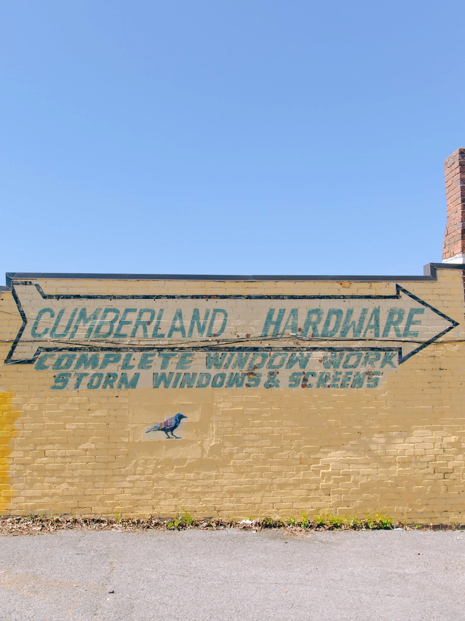 Brick wall with painted sign reading 'Cumberland Hardware' and additional text about windows and screen work, a small peacock illustration, and a clear blue sky above.