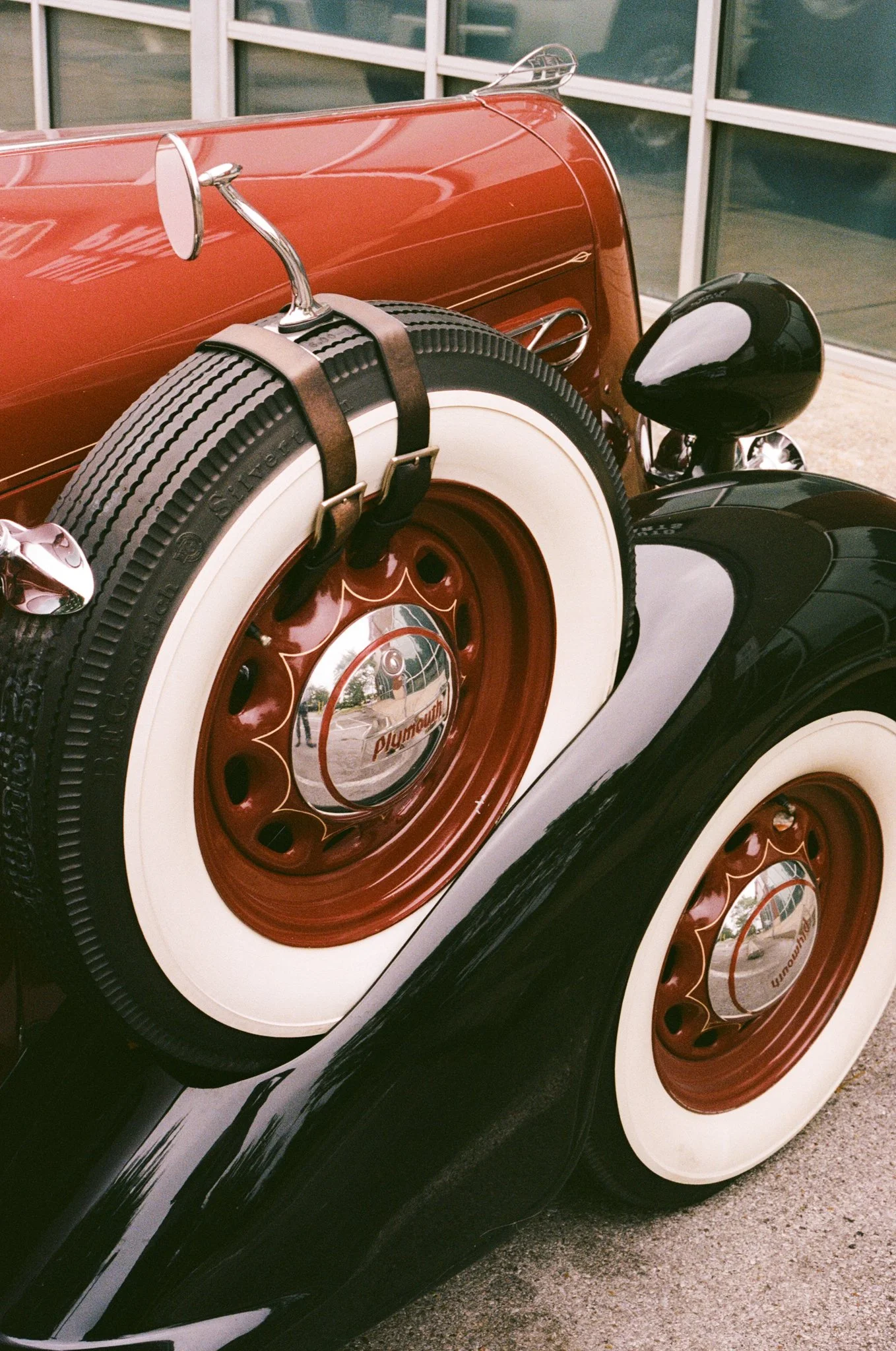 Close-up of the rear wheels of a vintage red and black automobile with whitewall tires and chrome hubcaps, parked outside near a building with glass windows.
