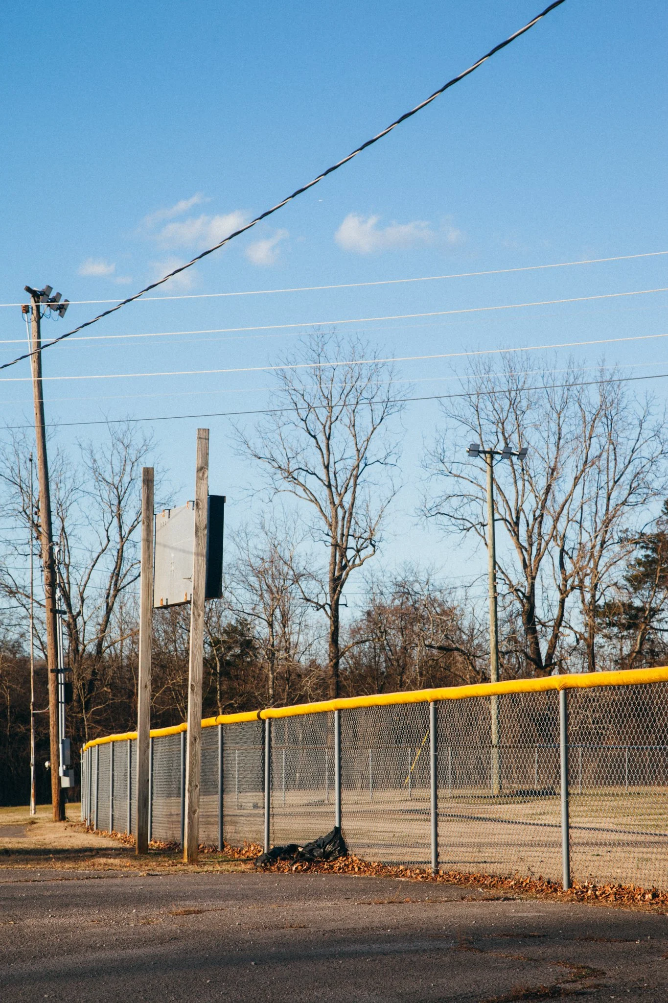 A baseball backstop fence with yellow top rail, several power poles with multiple wires, leafless trees, and a blue sky with a few clouds.