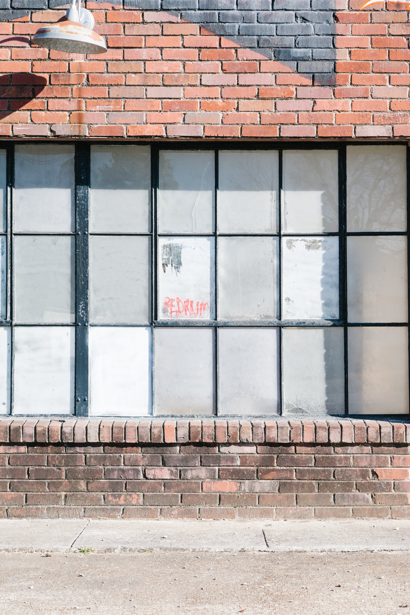 A brick building facade with a large window that has frosted glass panes and black metal framing. There is weathered graffiti in red spray paint on one of the window panes that says 'REDDRUM'. A vintage-style outside wall light fixture is mounted abo