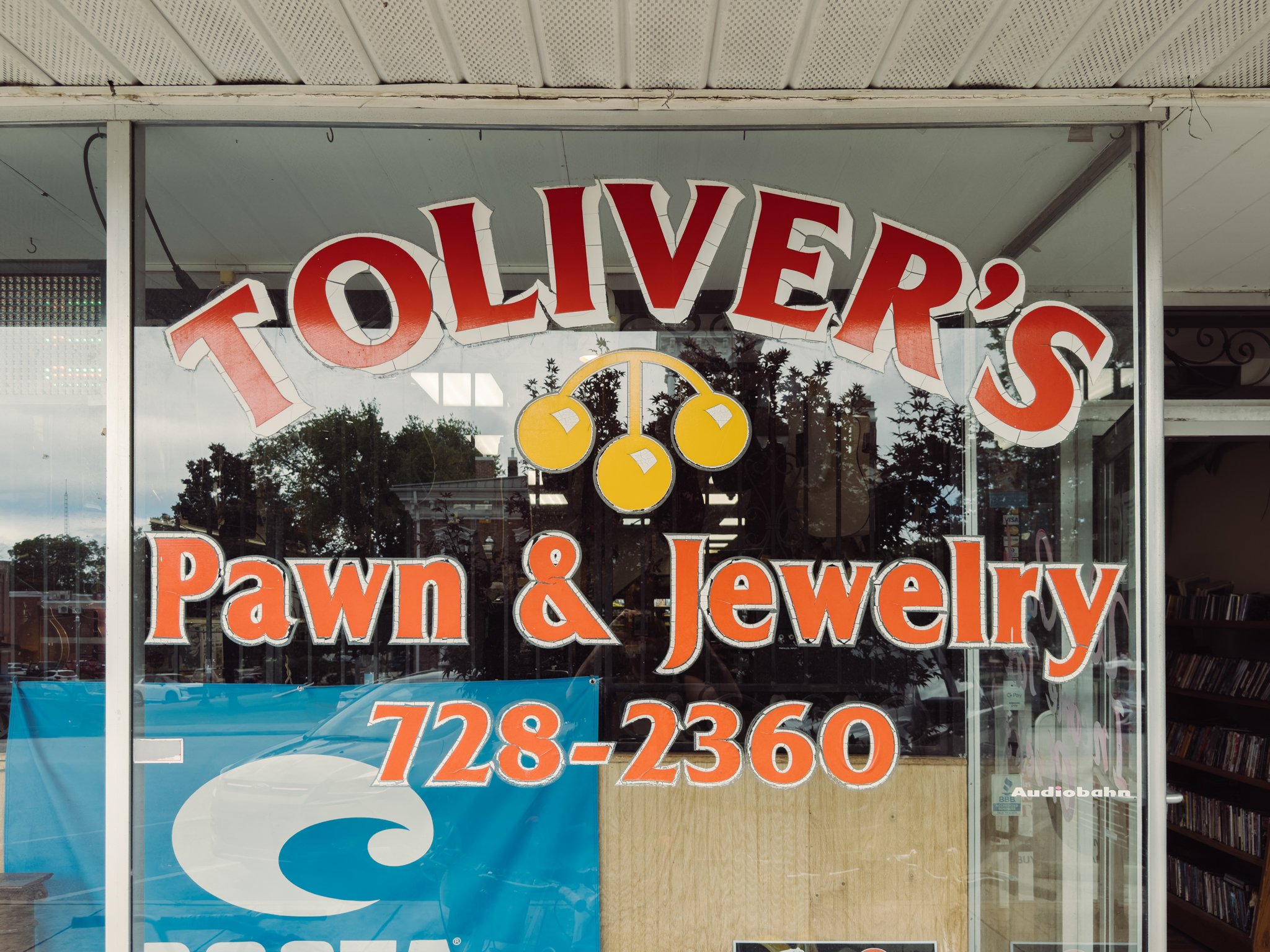 Storefront window with signs reading 'Oliver's Pawn & Jewelry' along with a phone number. The window reflects trees and the street outside.