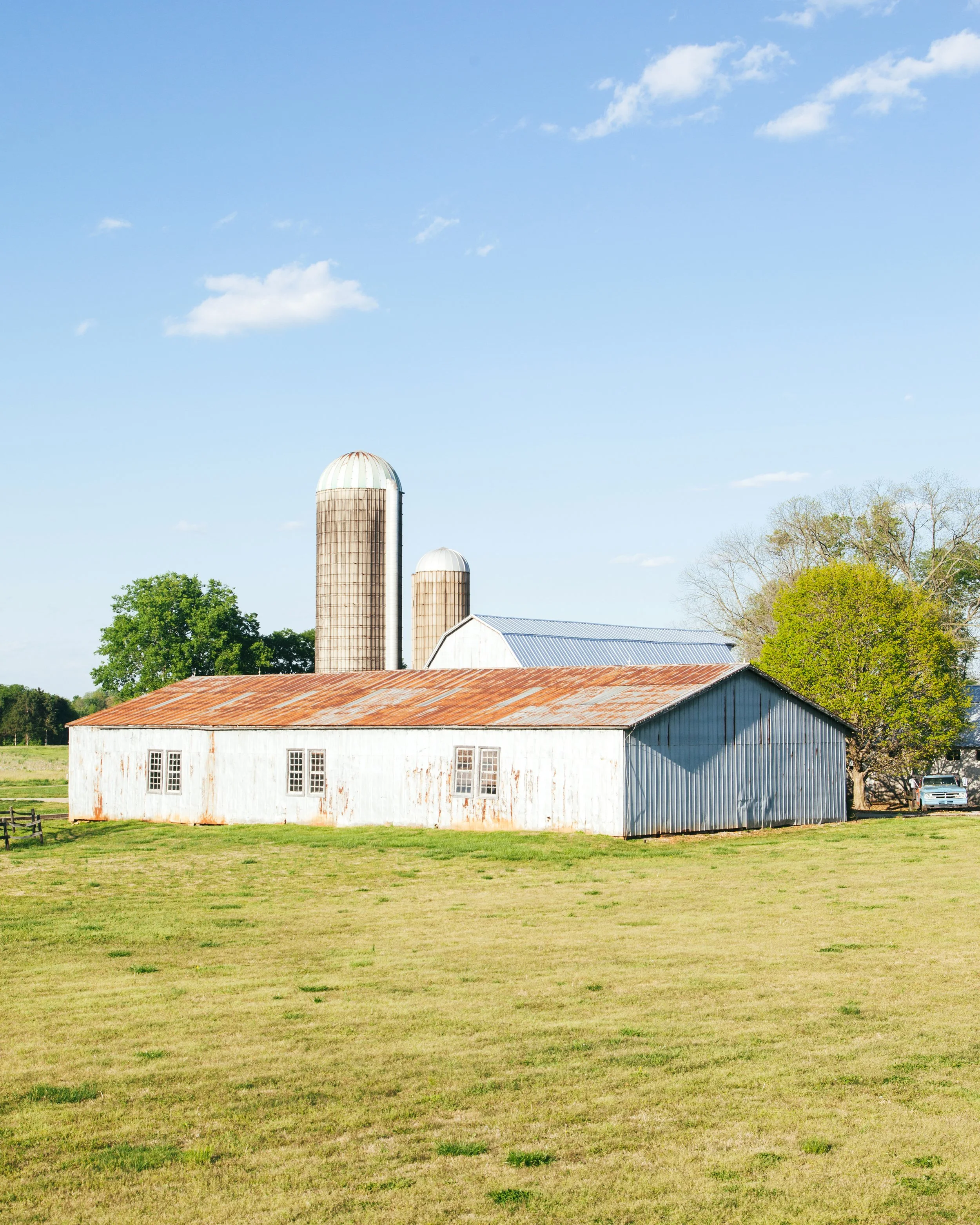 A rural farm scene with a large white barn, rust-colored roof, and three tall cylindrical silos. Green grass and trees surround the barn, with a blue sky and some clouds above.
