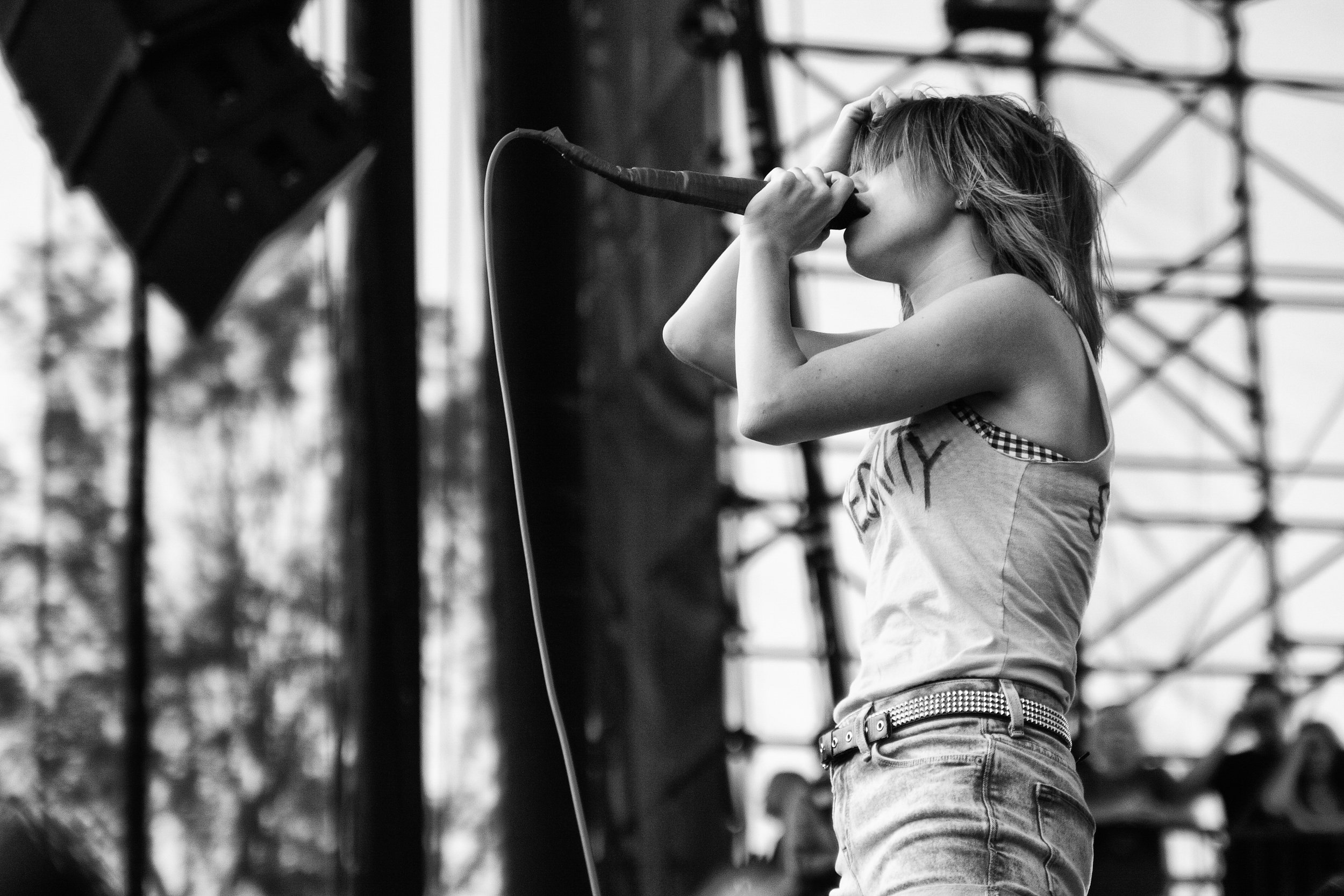 A young girl with blonde hair, wearing jeans and a sleeveless top, performs on stage with a microphone in her hand, appearing to sing or speak, with stage equipment and a scaffold in the background.