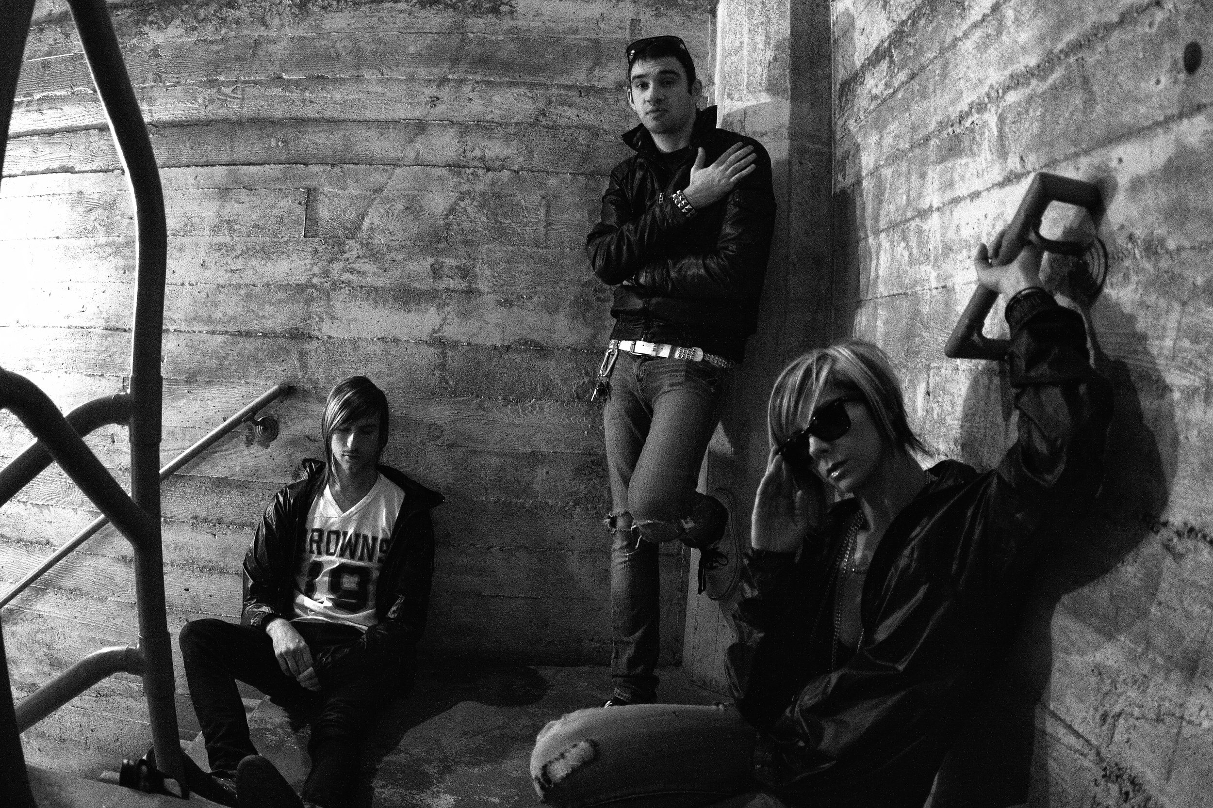 A group of four young adults with light skin posing against a rough concrete wall in an urban setting. They are wearing casual clothes, including jackets and sunglasses, and appear relaxed.