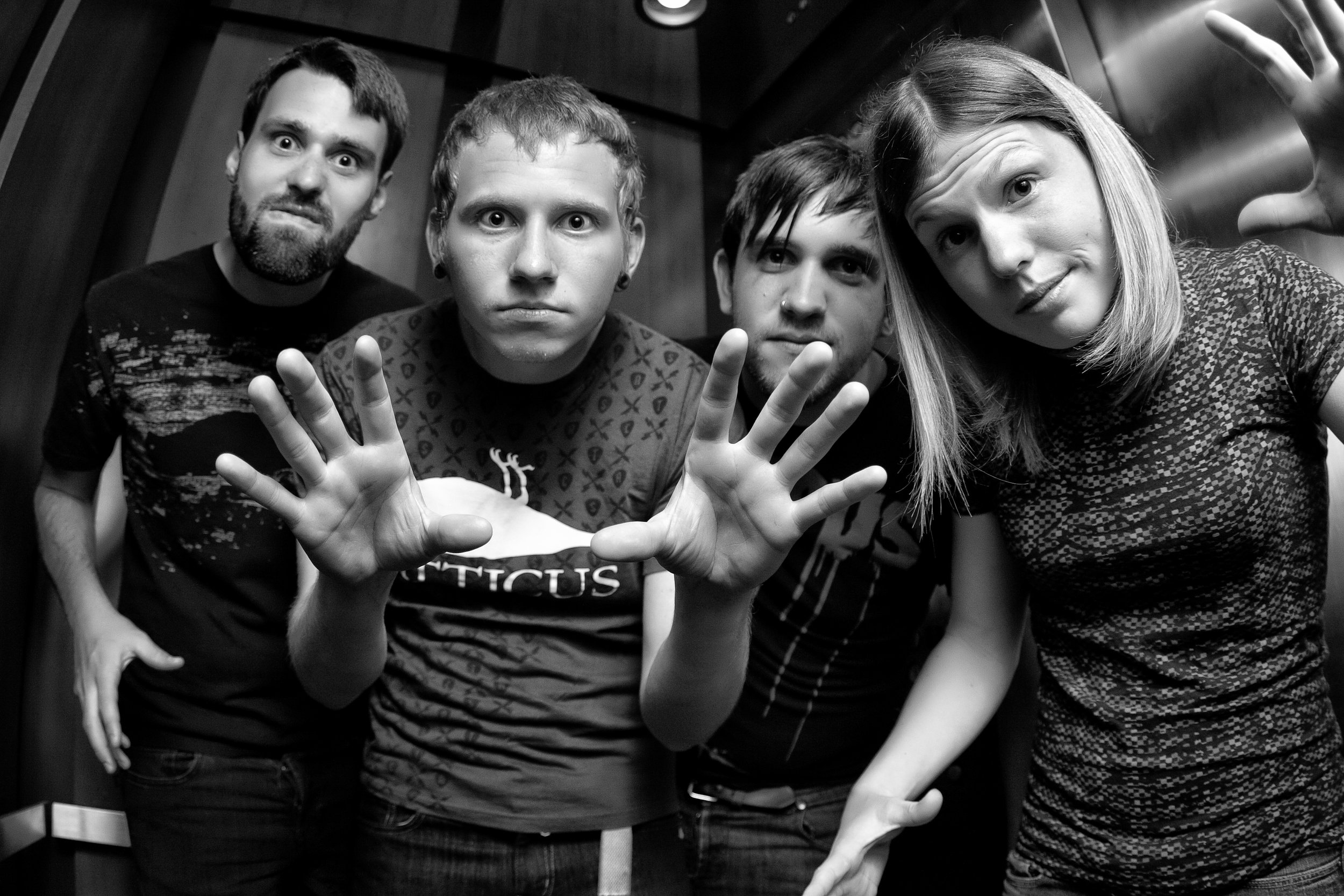 Four young adults, three men and one woman, posing inside an elevator. The woman and one man in front are making peace signs with their hands while the others look directly at the camera. The photo is in black and white.