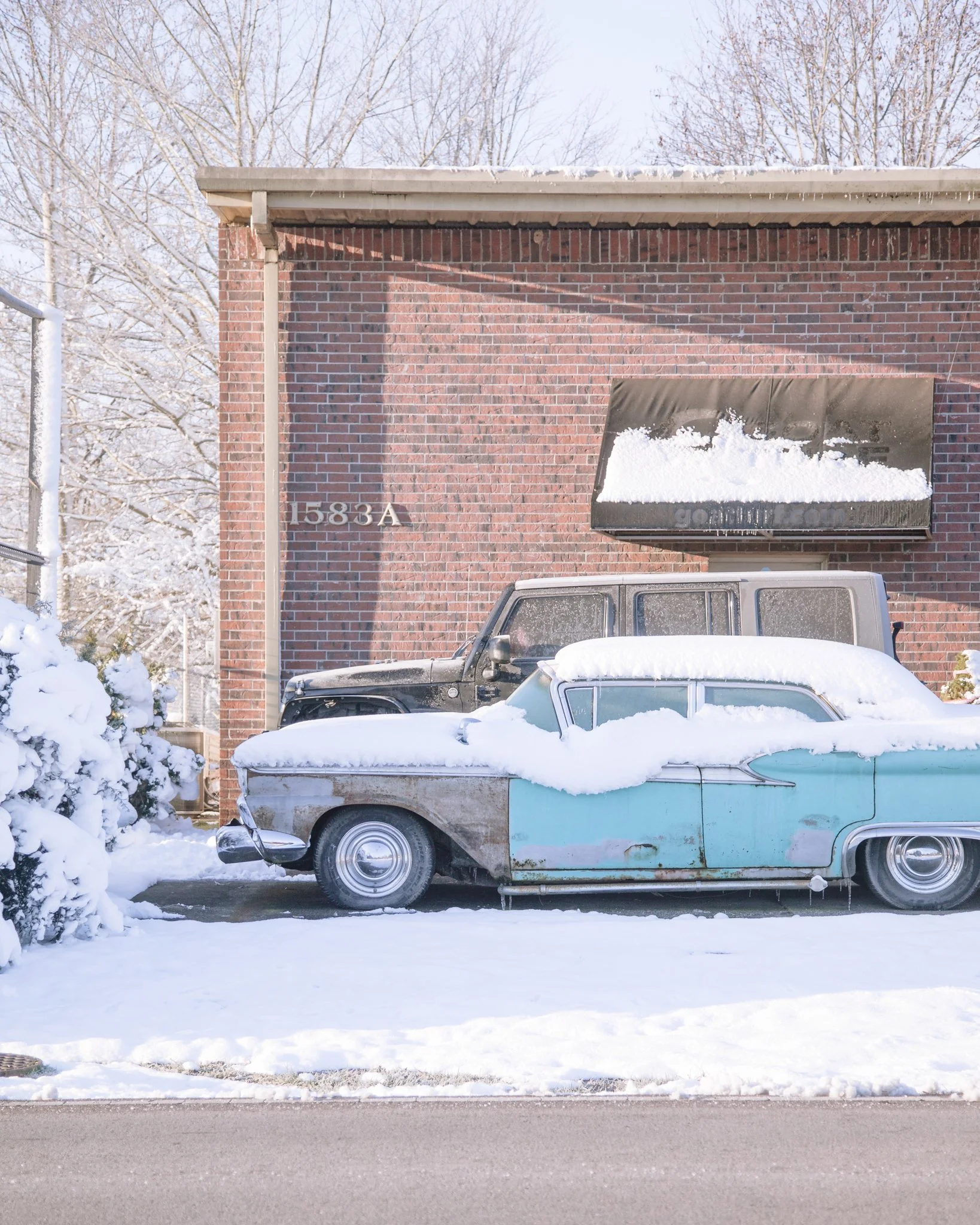 Snow-covered vintage car parked in front of a brick building with the address 1583A on it, during winter daytime.