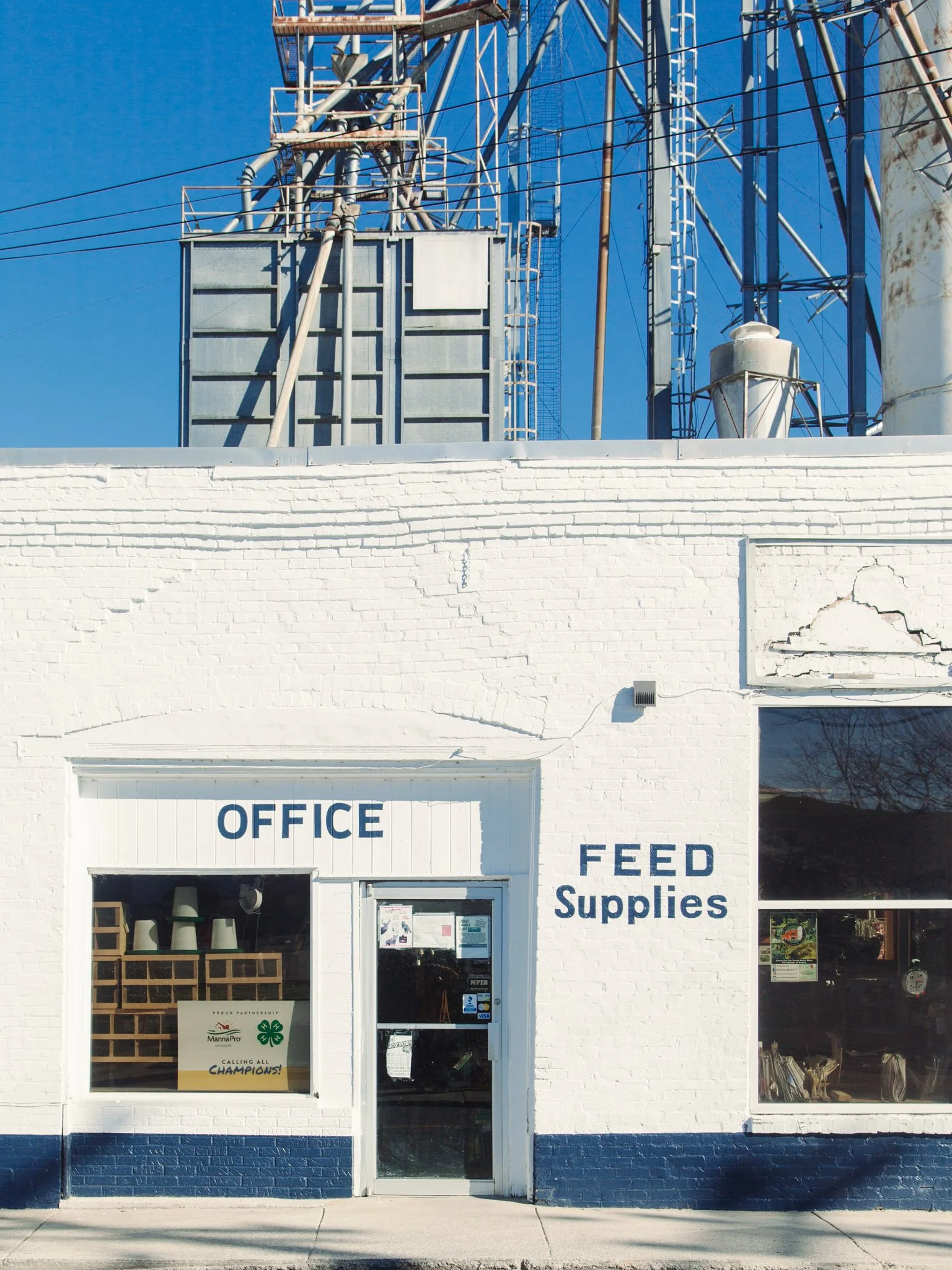 White building with signs reading 'Office' and 'Feed Supplies' on the front, glass door, large windows, and a white brick wall with some cracks, under a clear blue sky.