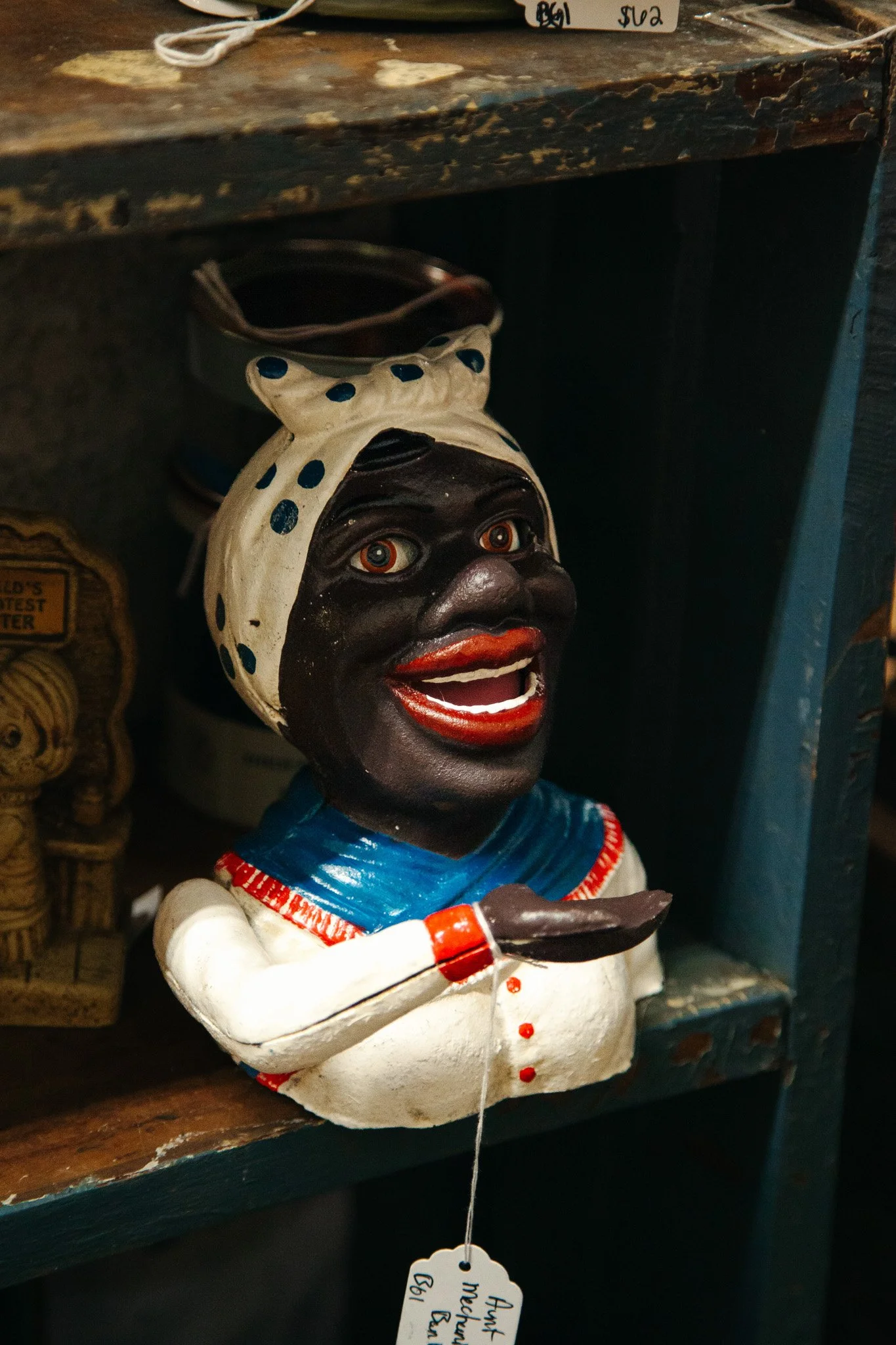 A decorative figurine of a smiling person with dark skin, painted face, and wide open eyes, wearing a white bonnet with blue polka dots and a blue and white outfit, holding a small object in one hand. The figurine is placed on a shelf with a handwrit