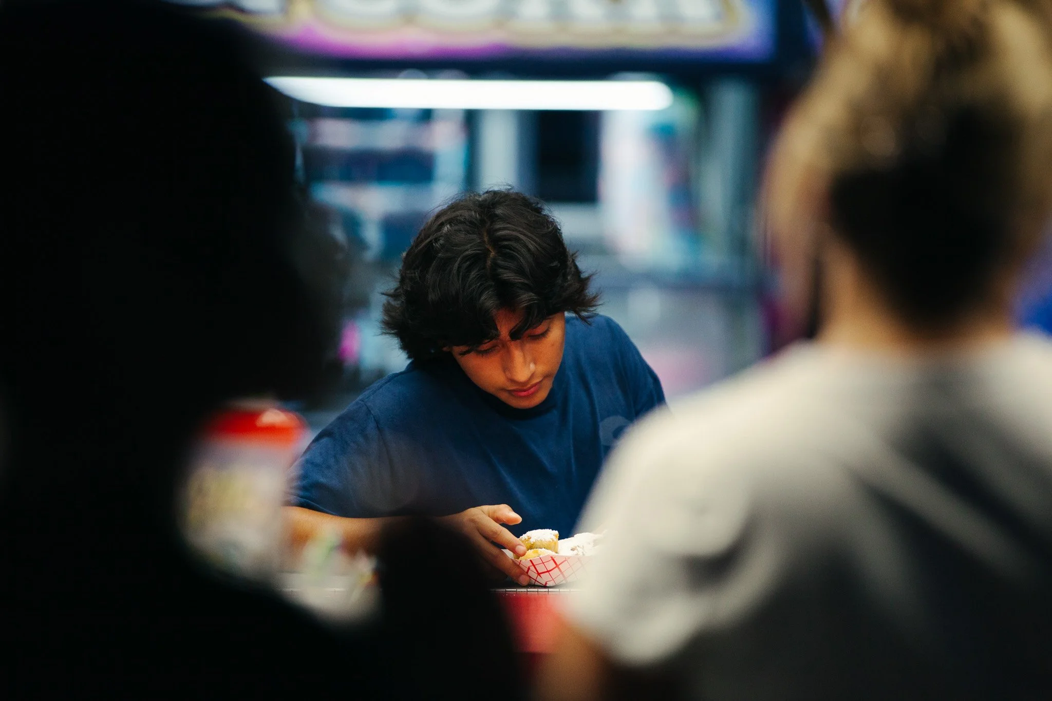 A young man with dark, curly hair is looking down while sitting at a table with a red and white checkered paper tray of food. The scene appears to be in a casual indoor setting, possibly a food stand or diner, with blurred figures and colorful lighti