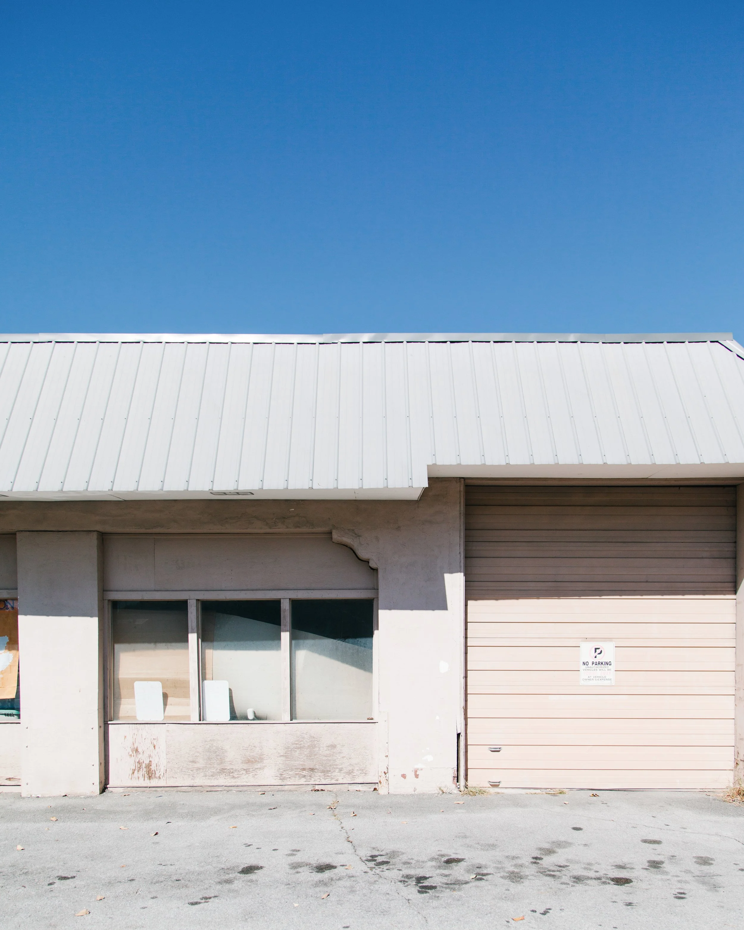 A storefront with a large, white, boarded-up window and a closed, beige roll-up door with a "No Parking" sign. The building has a white exterior with some paint chipping and a concrete sidewalk with scattered leaves and tire marks in front.