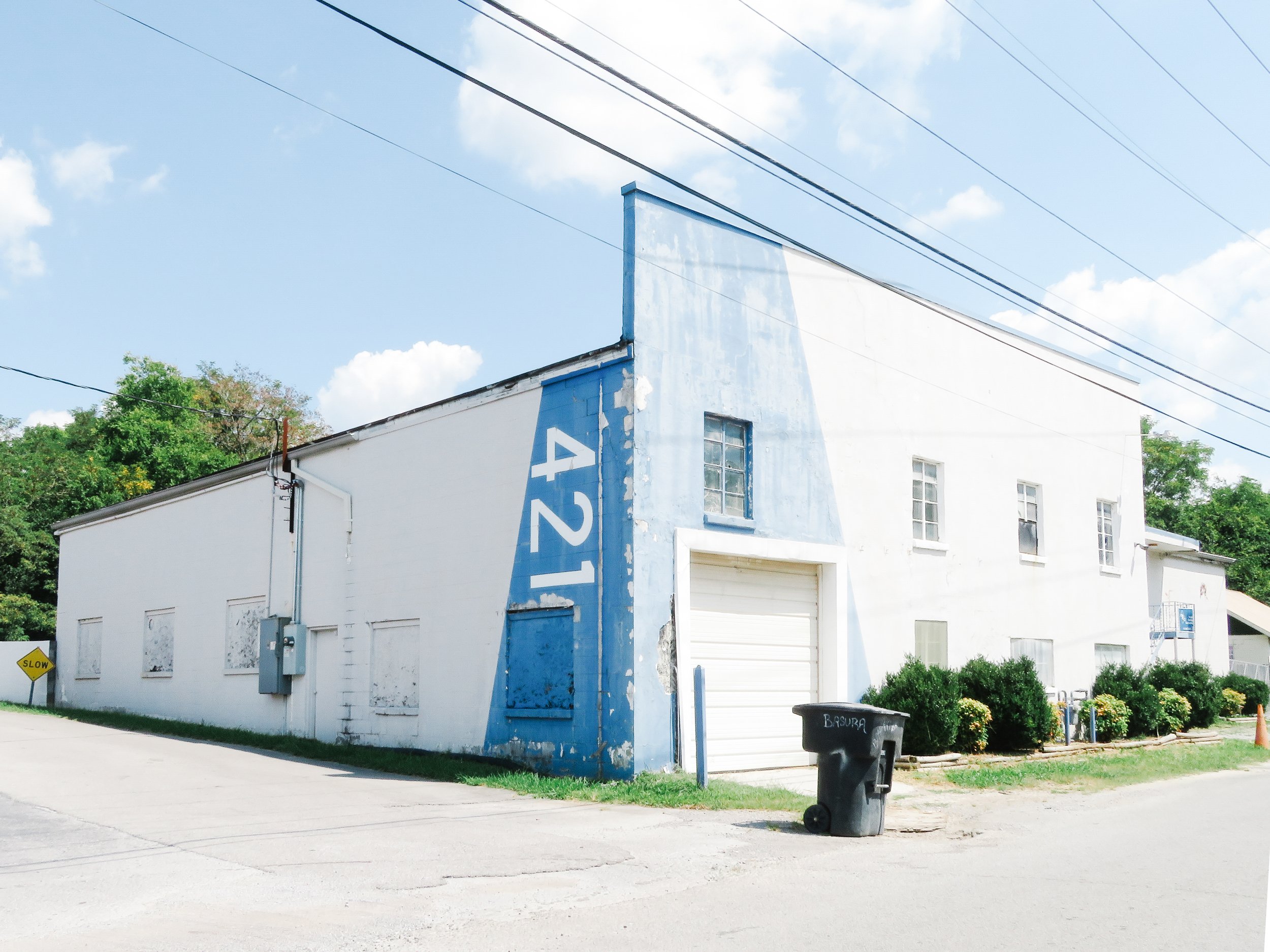 A white building with a blue painted section and a blue vertical sign that is faded. There is a small window on the side of the building. The area in front of the building includes a trash bin labeled "Basura," a garden with bushes and flowers, a yel