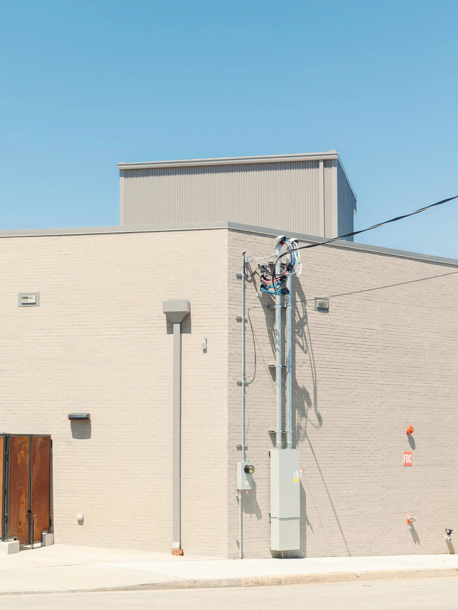 Side view of a beige brick building with electrical installations, utility boxes, and a fire safety sign, under a clear blue sky.