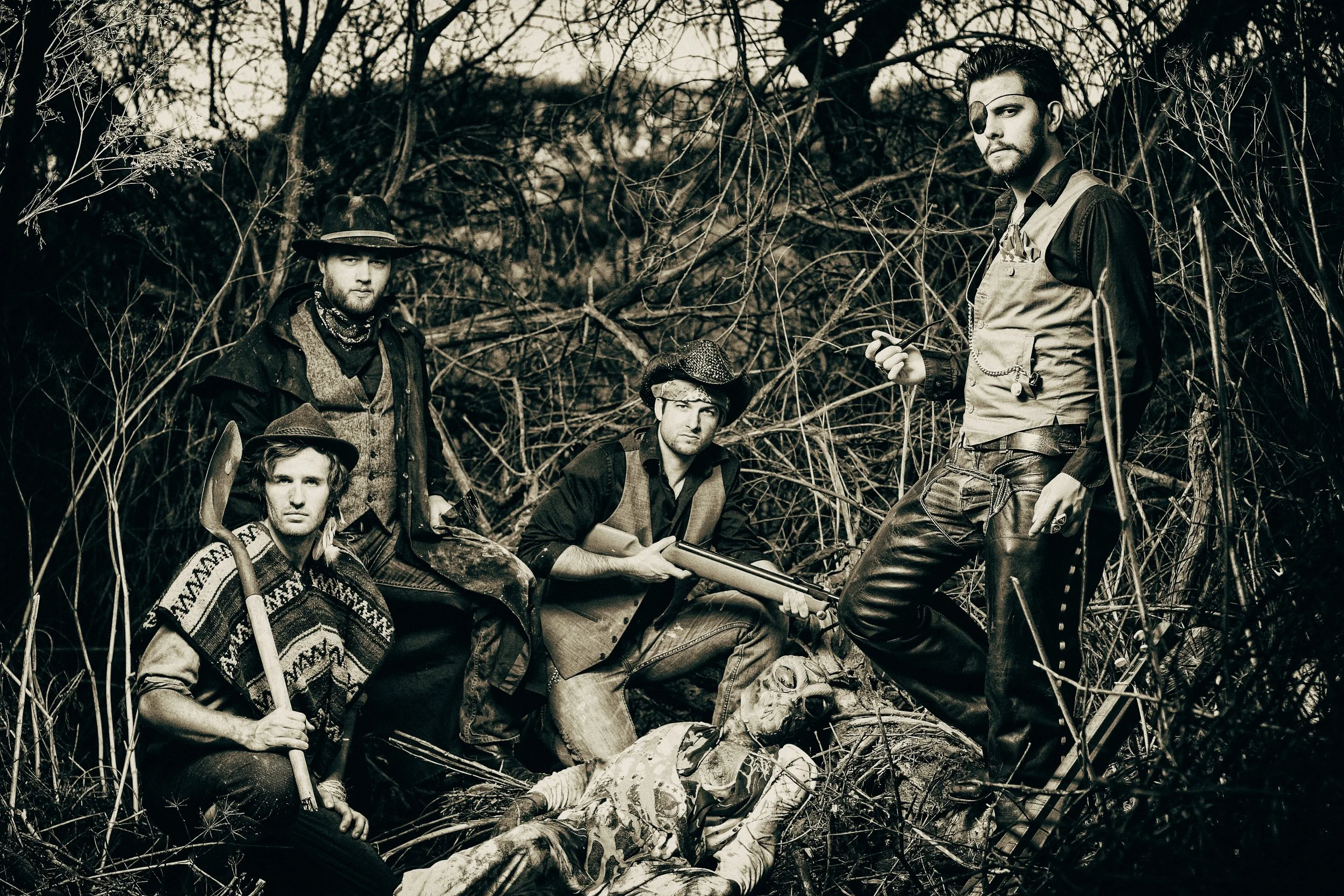 A group of five individuals dressed as hunters in a wooded area with leafless trees, wearing vintage or rustic clothing, some with hats, and holding tools or weapons, posing for a photo in a monochrome style.