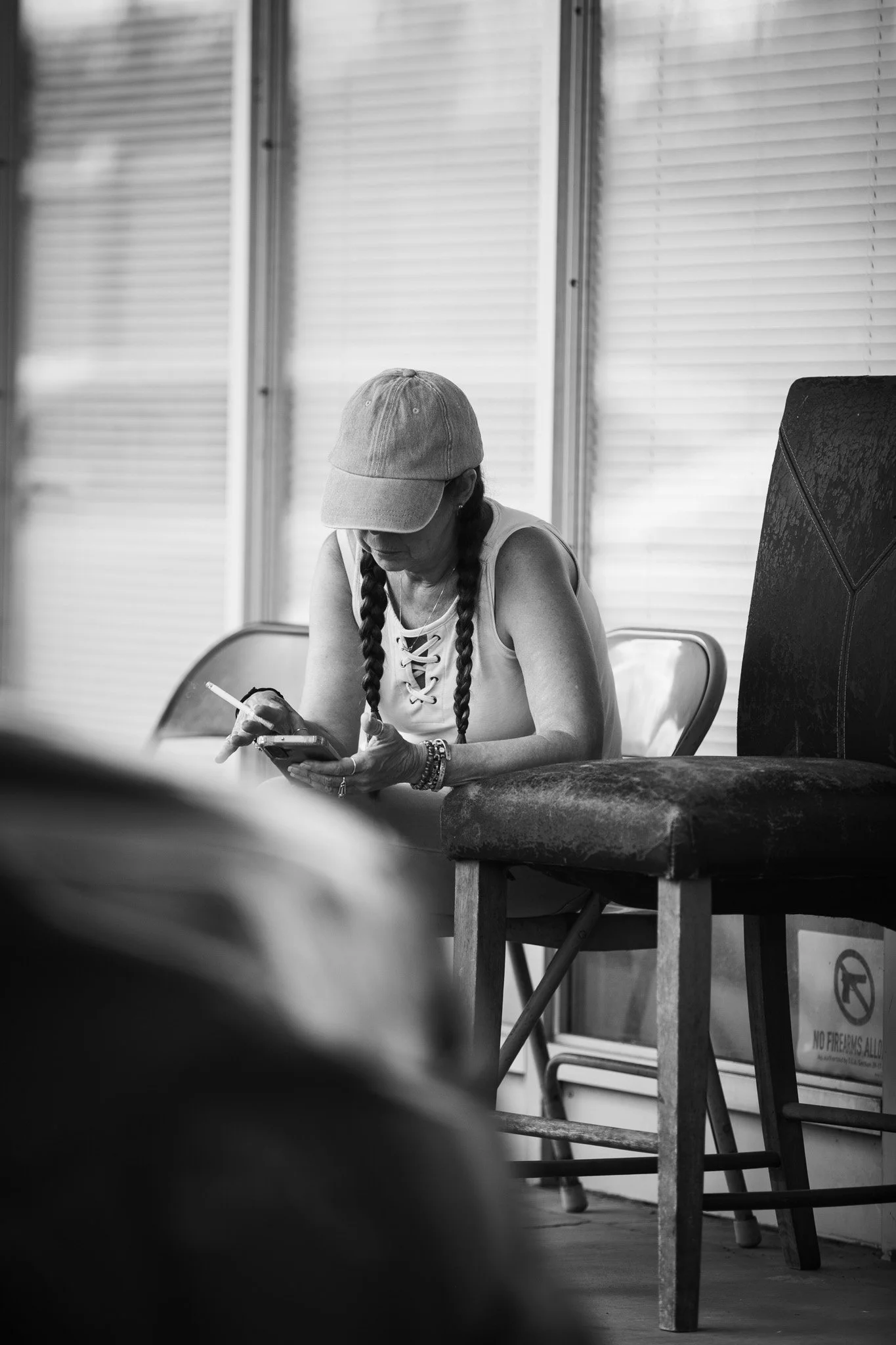 A woman wearing a cap and braided hair is sitting on a chair, looking at her phone, in front of window blinds.
