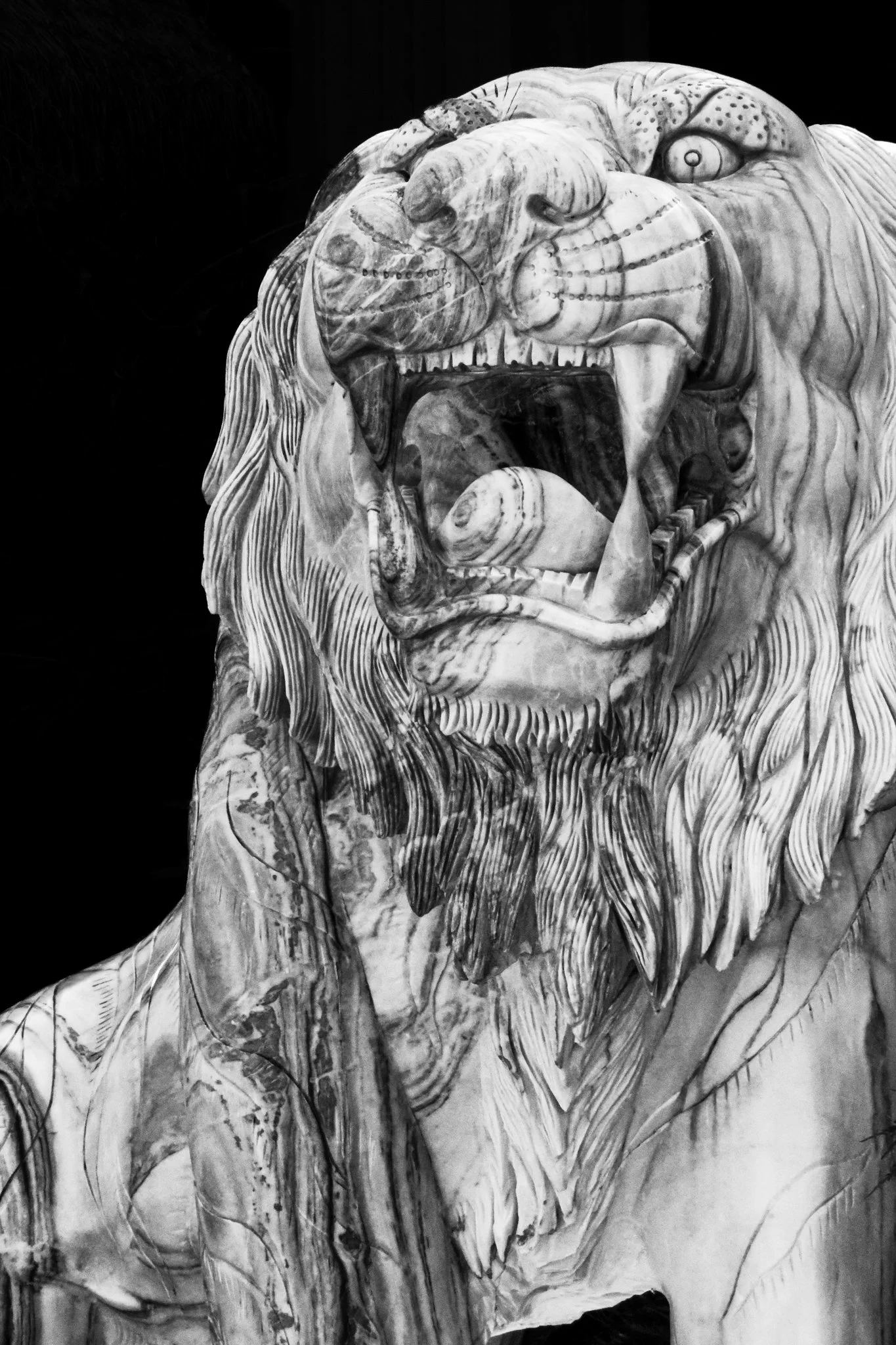 A carved sculpture of a roaring lion with detailed features, including a mane, open mouth with visible teeth and tongue, and expressive eyes, set against a dark background.