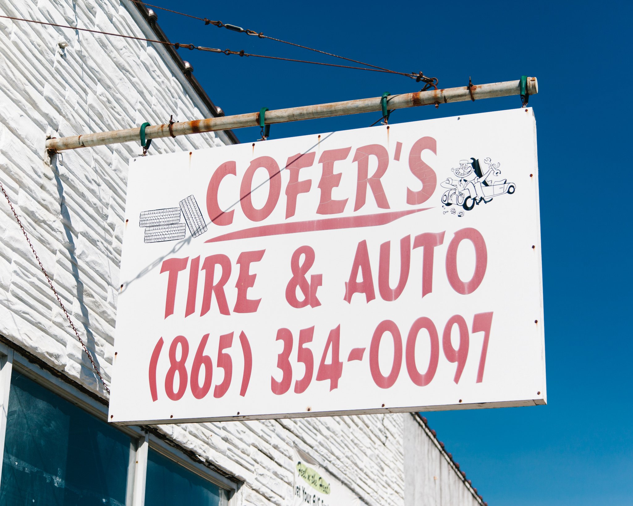 A signboard hangs outside a building with a white brick exterior, advertising an auto shop for tires and auto services, with a phone number.