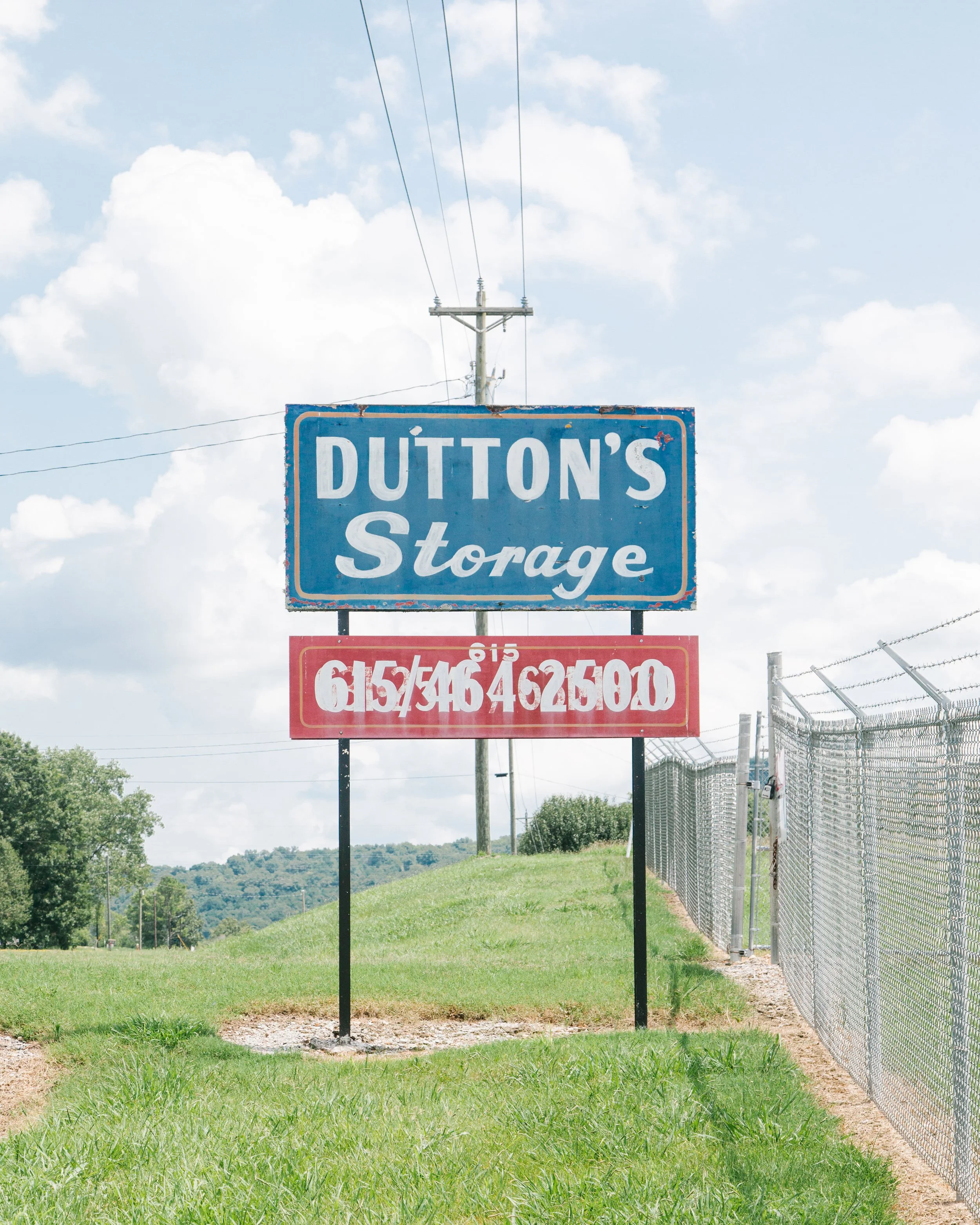 A blue sign with white text reading 'Dutton's Storage' is hanging from a pole in front of an open grassy area with trees and rolling hills in the background. The sky is mostly cloudy with some blue showing, and a fence runs along beside the sign on t