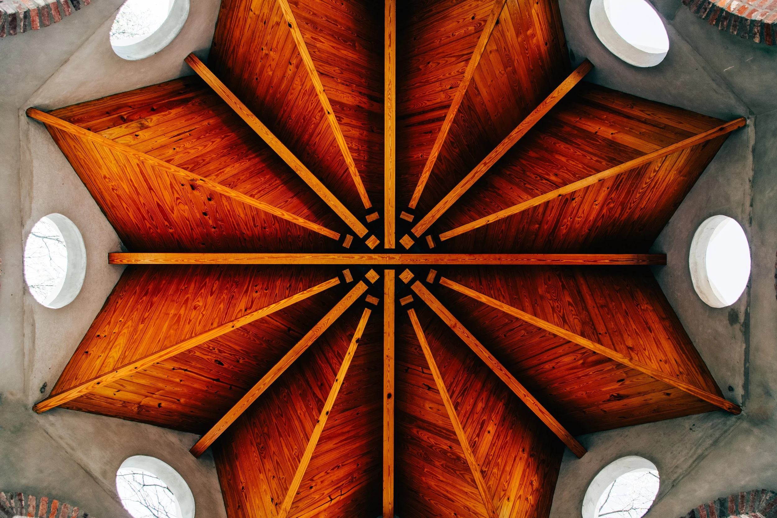 View of a wooden ceiling with a geometric pattern, surrounded by circular windows in a concrete structure.