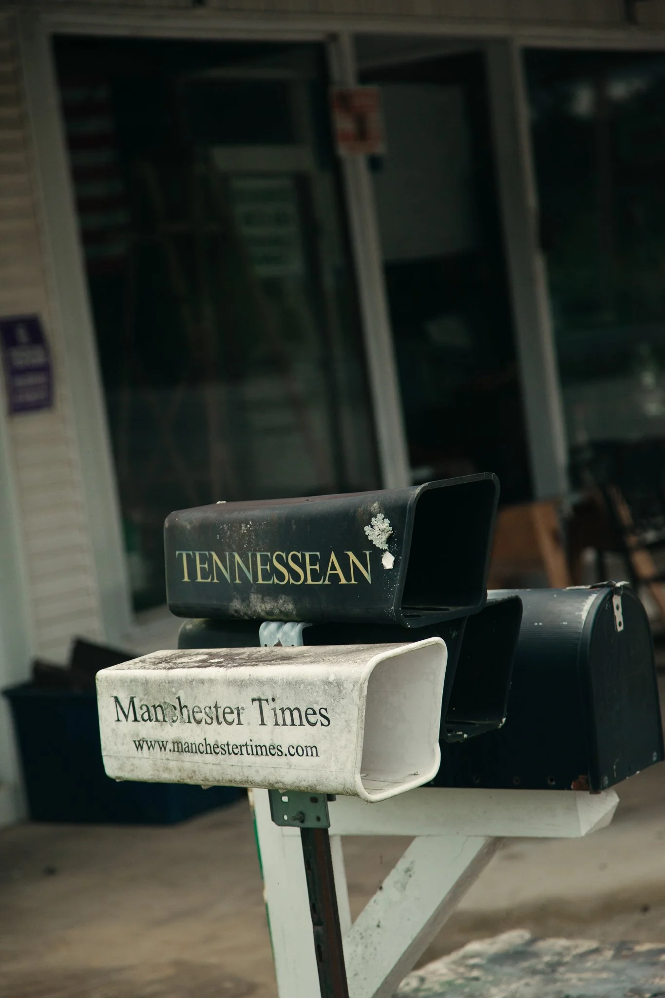 Close-up of a mailbox with a Tennessee State flag sticker and a Mancherster Times newspaper. In the background, there are glass-fronted display cases with various items inside, outside a building.