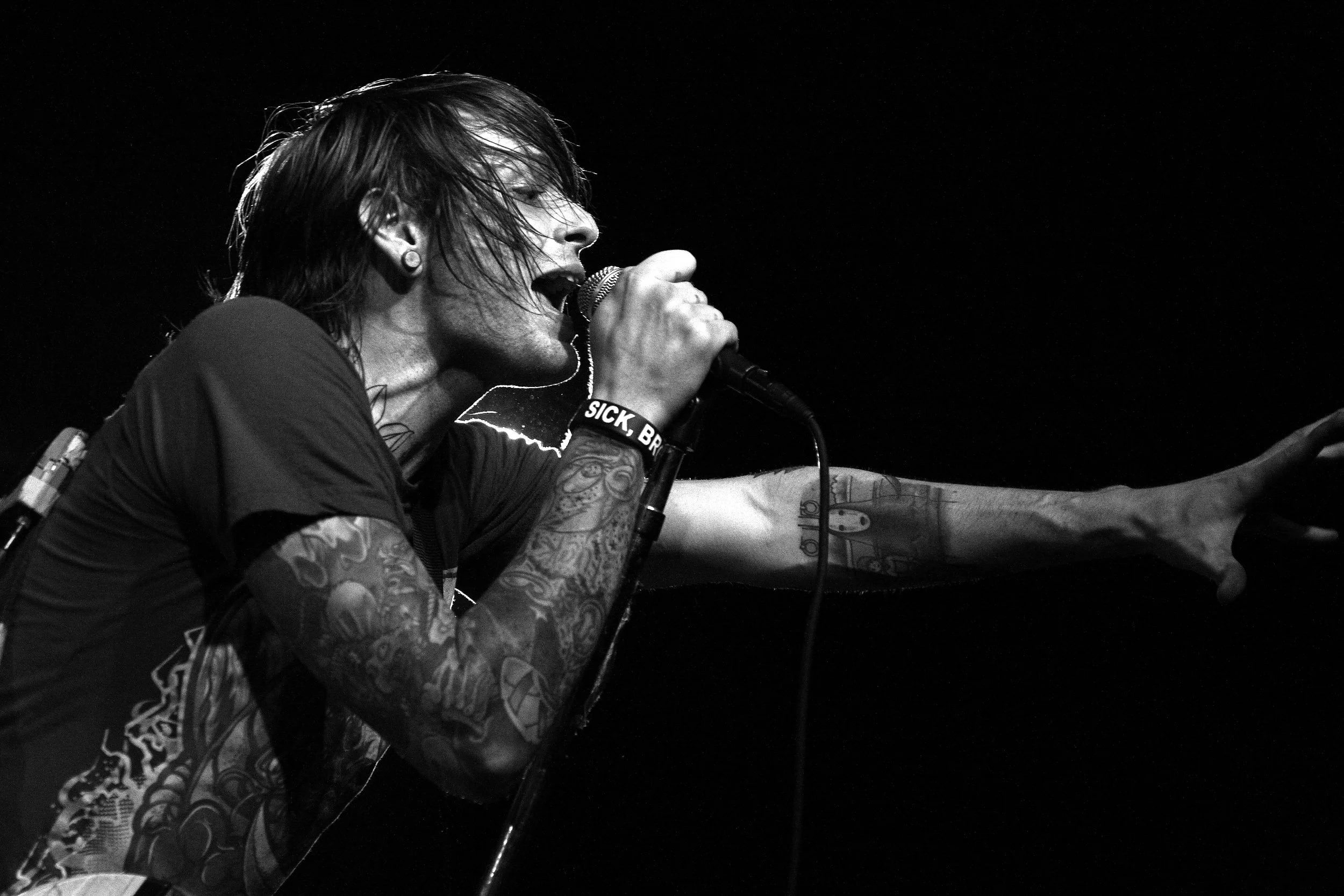 Black and white photo of a person singing into a microphone. They have long dark hair, visible tattoos on their arm, and are wearing a wristband that says 'SICK, BRAVE'.