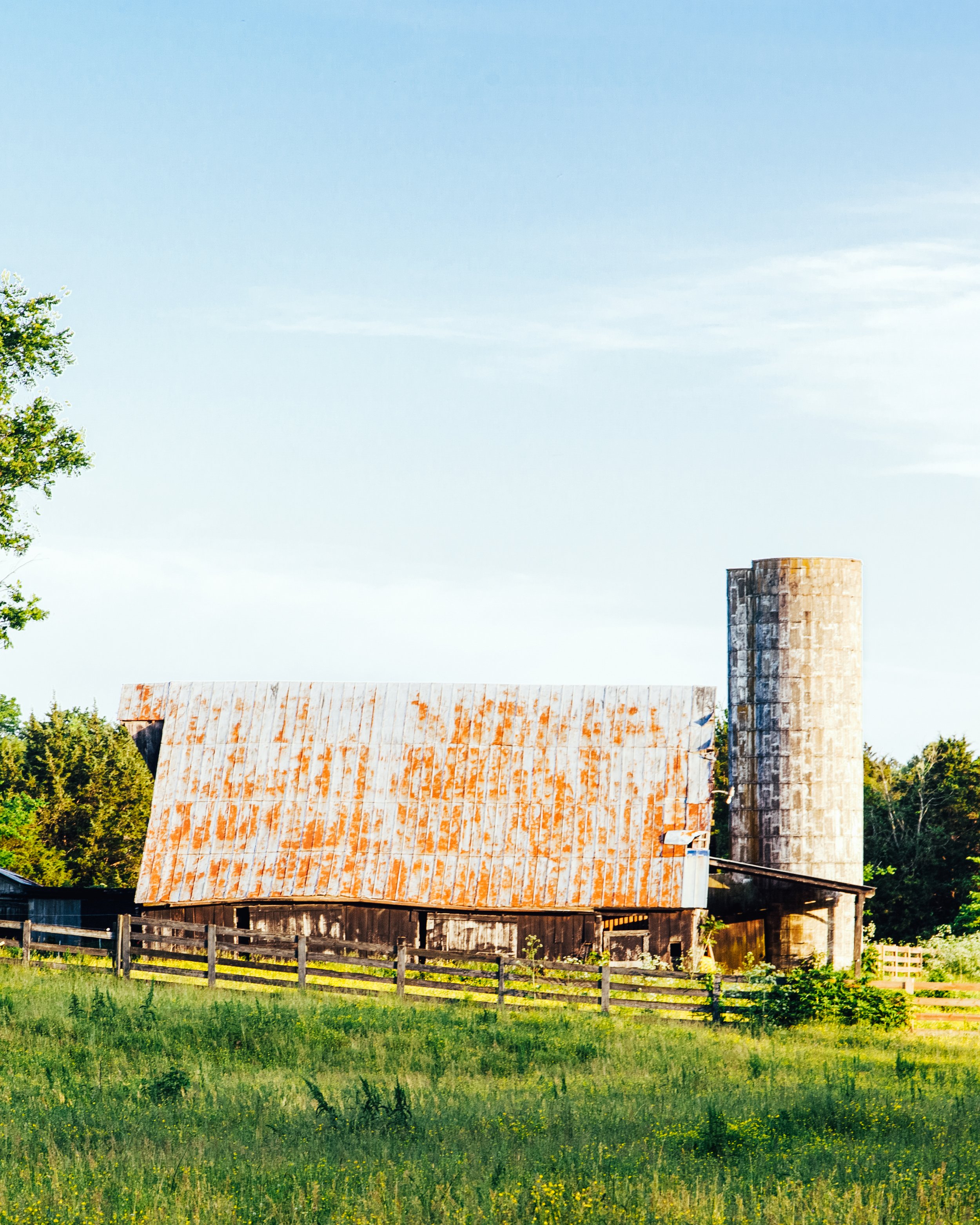 An old barn with rusted metal roof, a chimney, surrounded by green grass, trees, and a wooden fence under a blue sky.