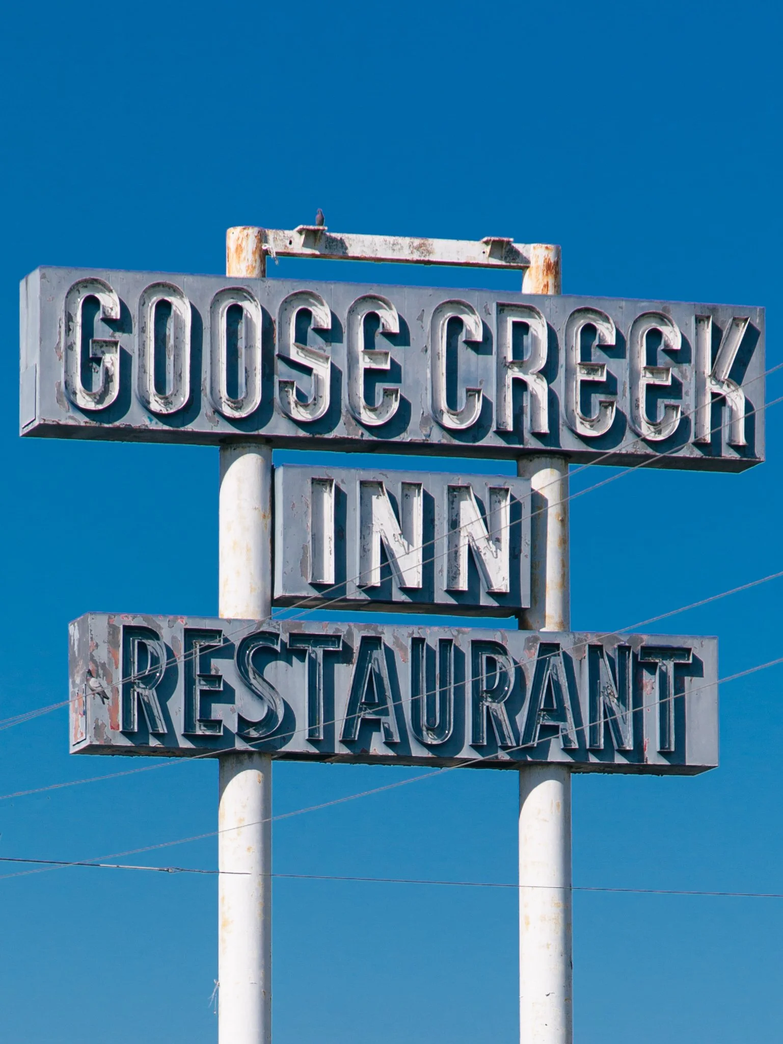 An old, weathered sign for a business named 'GOOSE CREEK' on a clear blue sky background. The sign is rusted and faded, and the text is in large capital letters.
