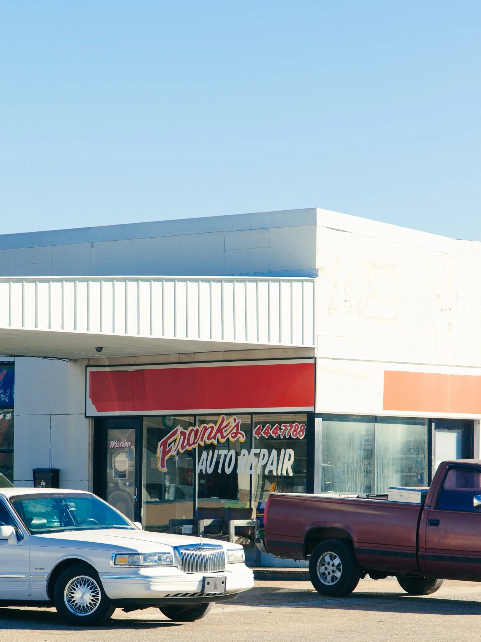 A storefront with a sign that says 'Frank's Auto Repair.' There are two cars parked in front: a white sedan and a red pickup truck, under a light blue sky.