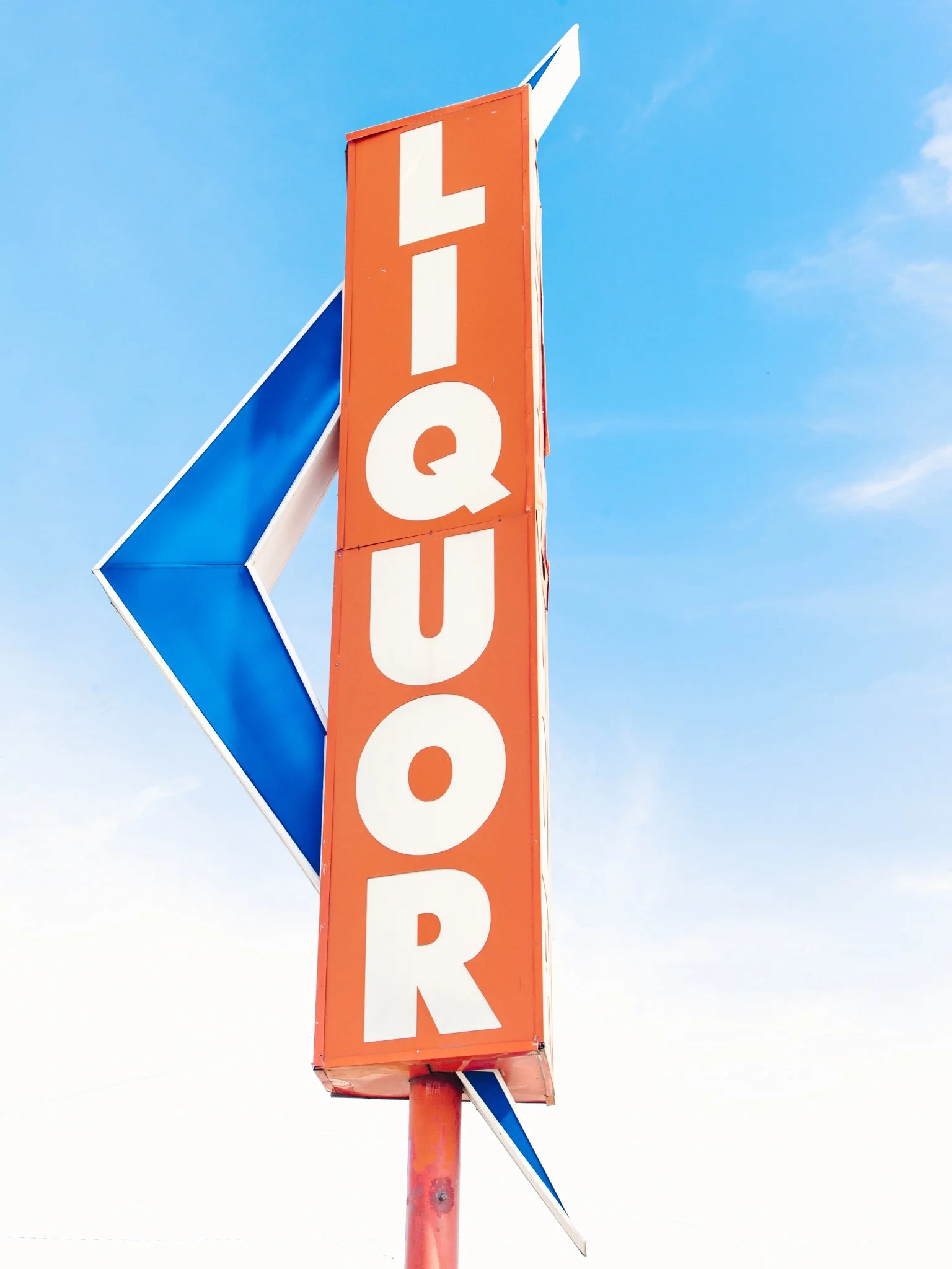 A tall orange sign with the word 'LIQUOR' in white letters, attached to a pole, with a blue and white arrow-shaped sign below pointing to the left, against a clear blue sky.