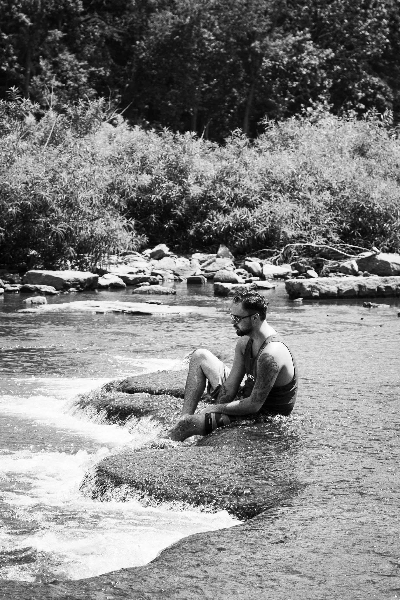 A man with tattoos and glasses sitting on rocks at the edge of a river with trees and bushes in the background.