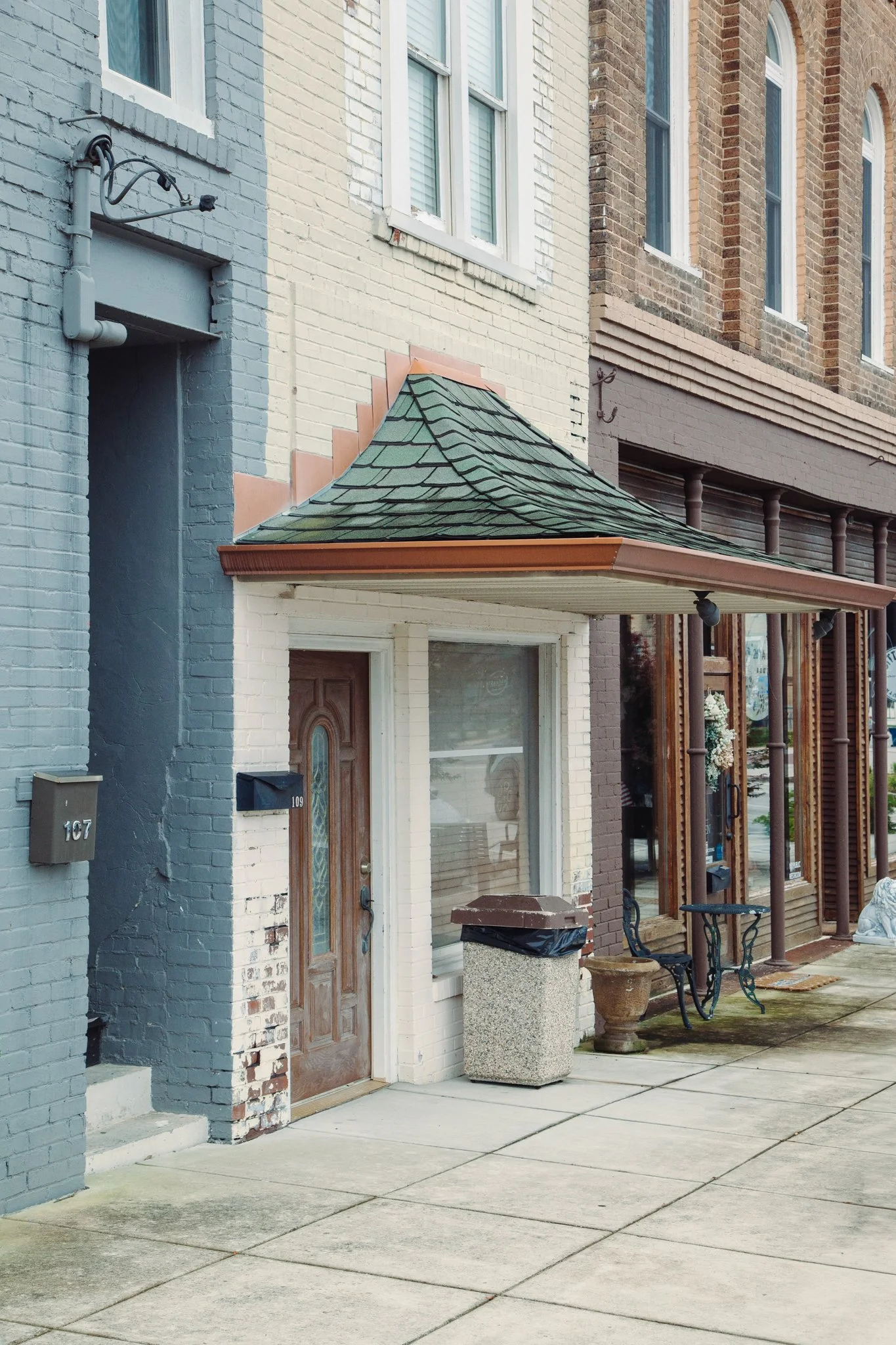 Three adjacent buildings with brick facades, each with a distinct paint color, with storefronts and address numbers 107 and 109 on the sidewalk. A small overhang roof with green shingles is over the entrance of building 109, and in front of it is a l