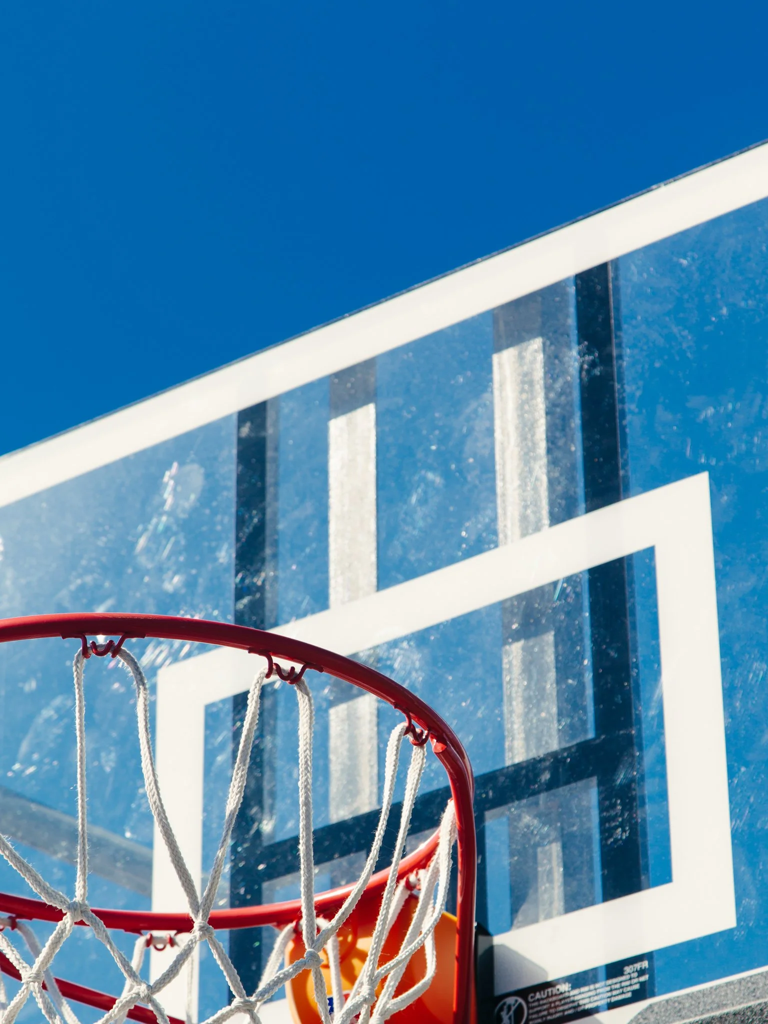 A view from beneath a basketball hoop looking upward at a blue sky with white clouds. The basketball backboard and hoop are visible, with a ball passing through the net.