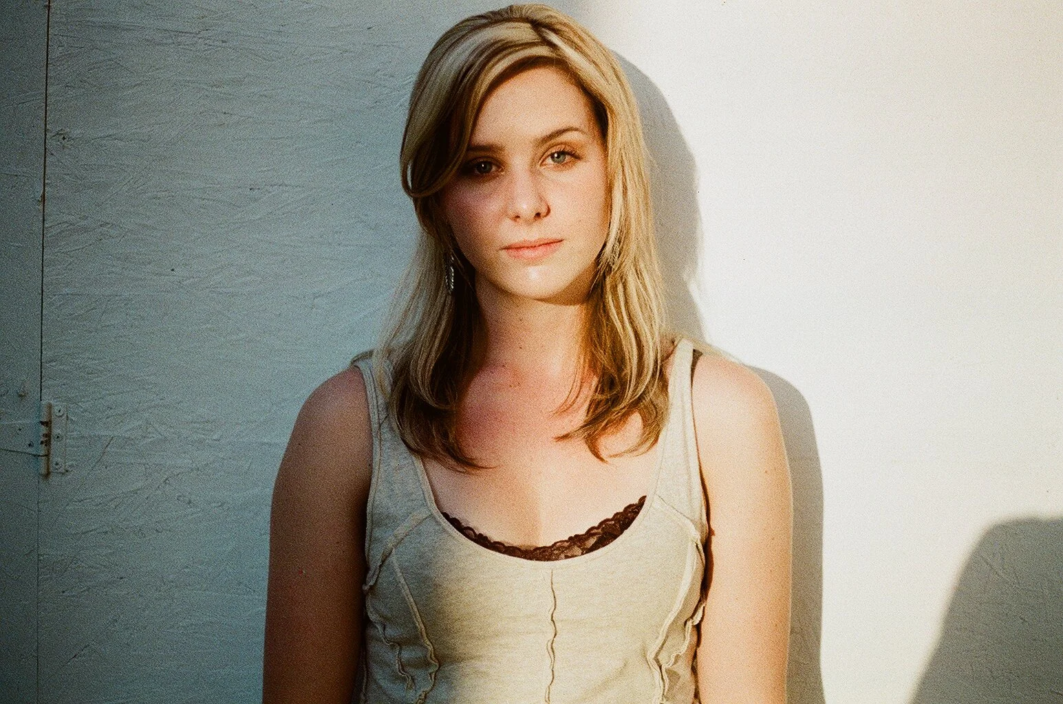 A young woman with blonde hair and light skin standing against a textured wall, wearing a tank top with lace detail.
