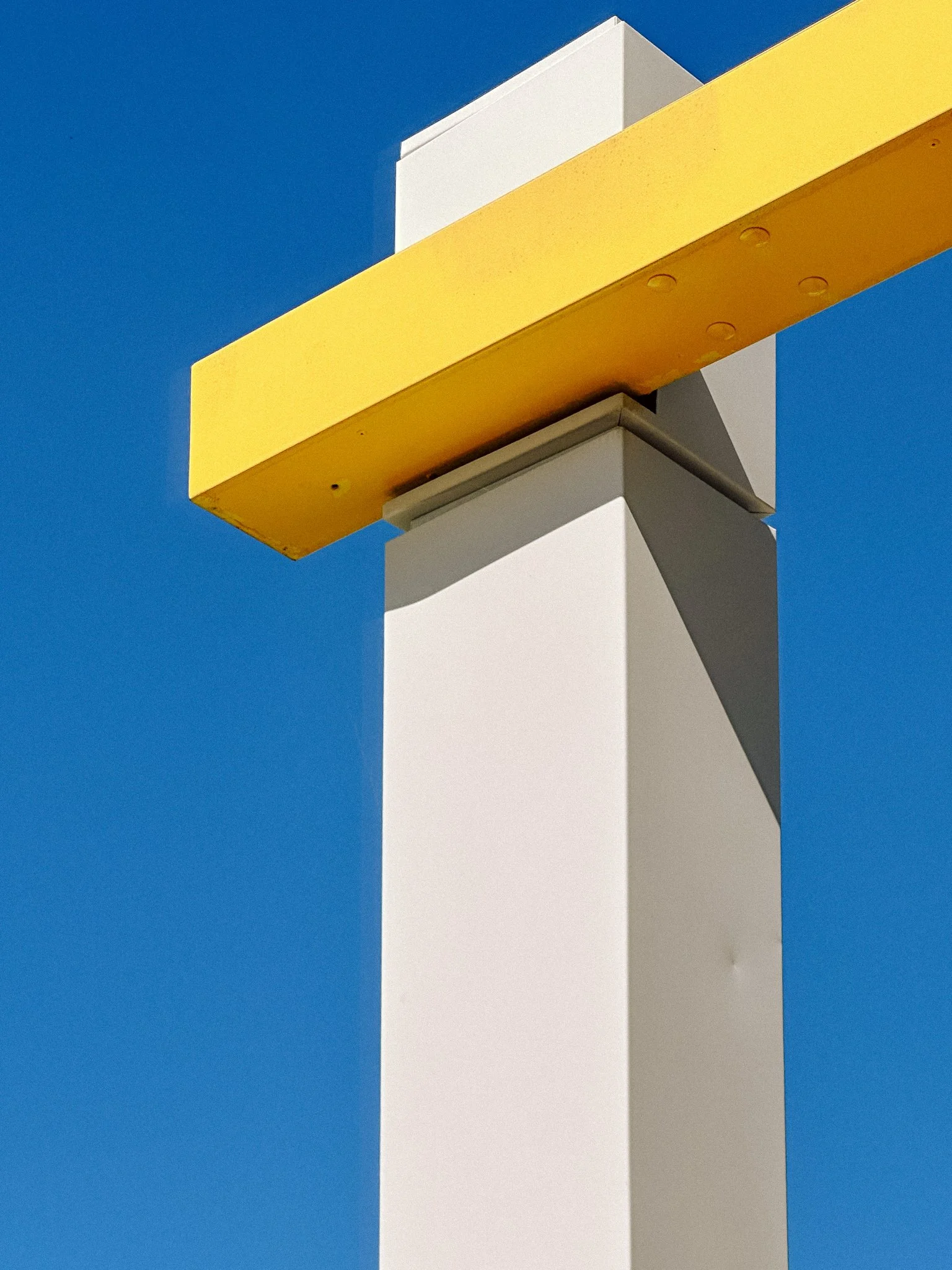 Close-up of a modern street light post against a clear blue sky, featuring a white pole and a yellow horizontal arm extending from it.