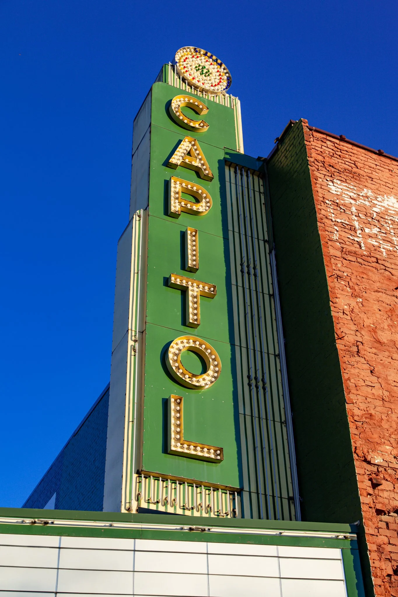 Vintage neon sign for a hotel, with large yellow letters outlined with light bulbs, attached to a tall green building against a clear blue sky, with part of a brick building visible on the right.