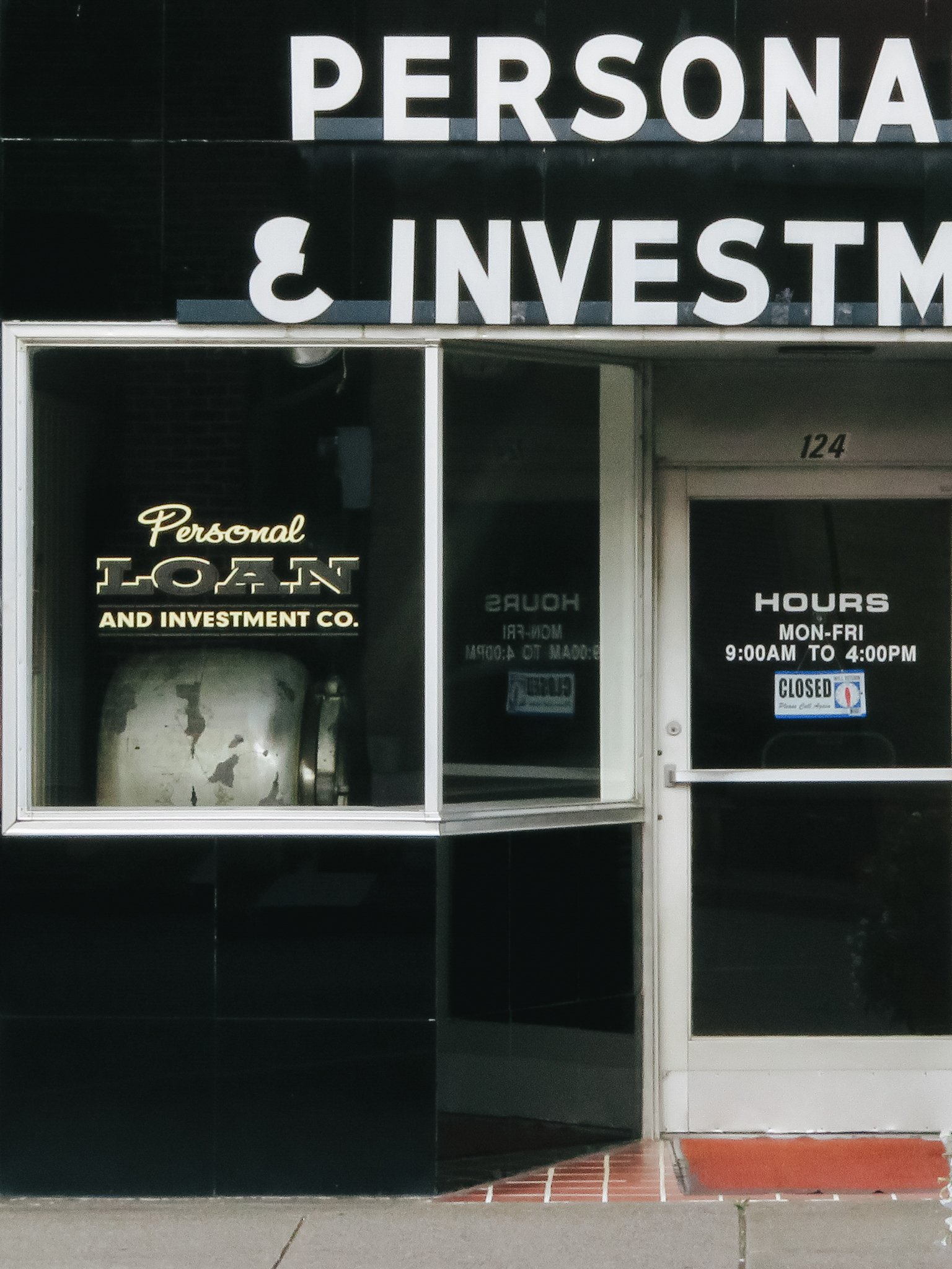 Storefront with black and white exterior signage that reads 'PERSONAL & INVESTMENT' with the store hours listed as Monday to Friday 9:00 AM to 4:00 PM and a closed sign in the window.