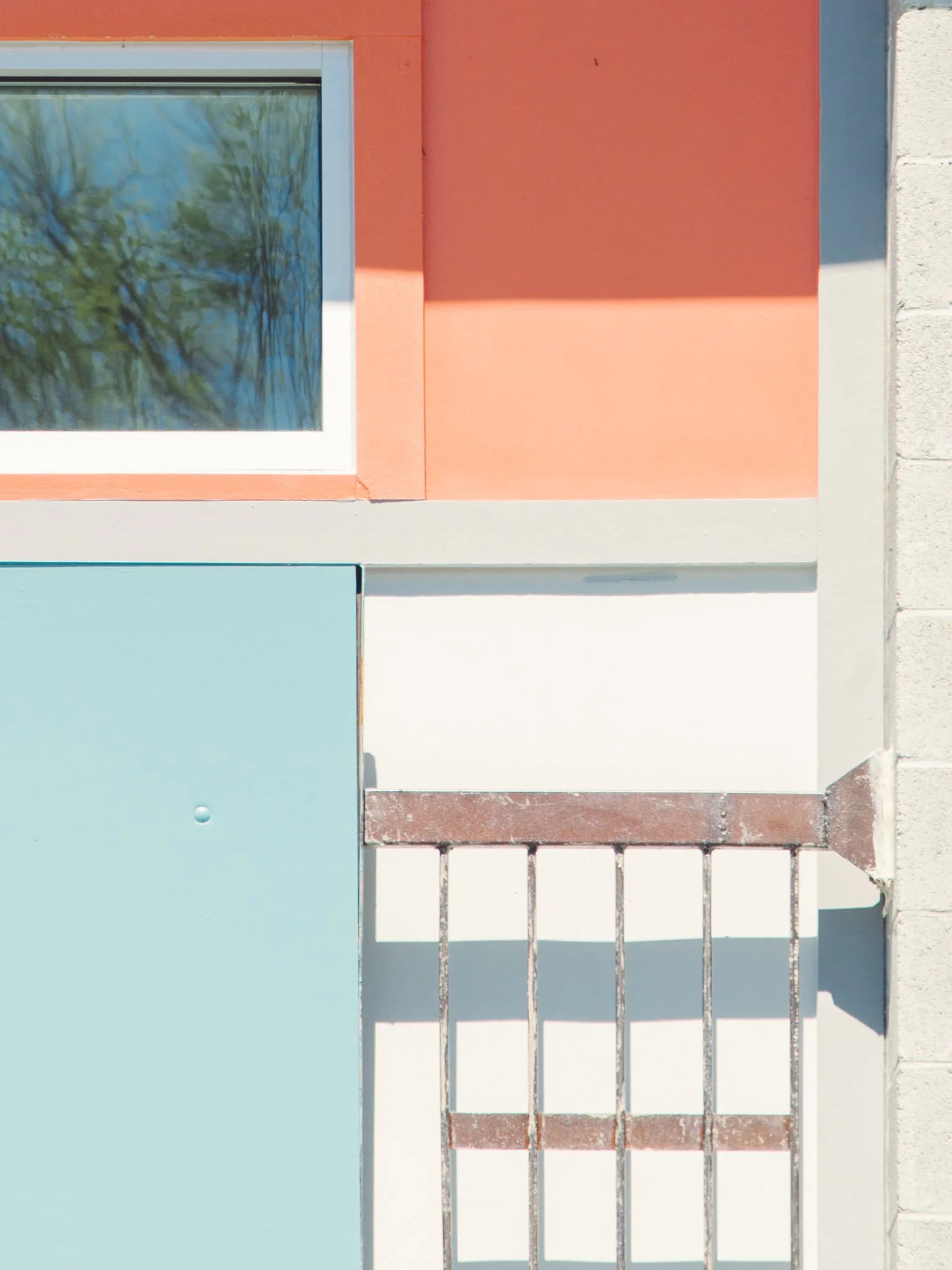 Close-up of a colorful modern building exterior with a window reflecting trees, orange and blue panels, white trim, and a metal railing.