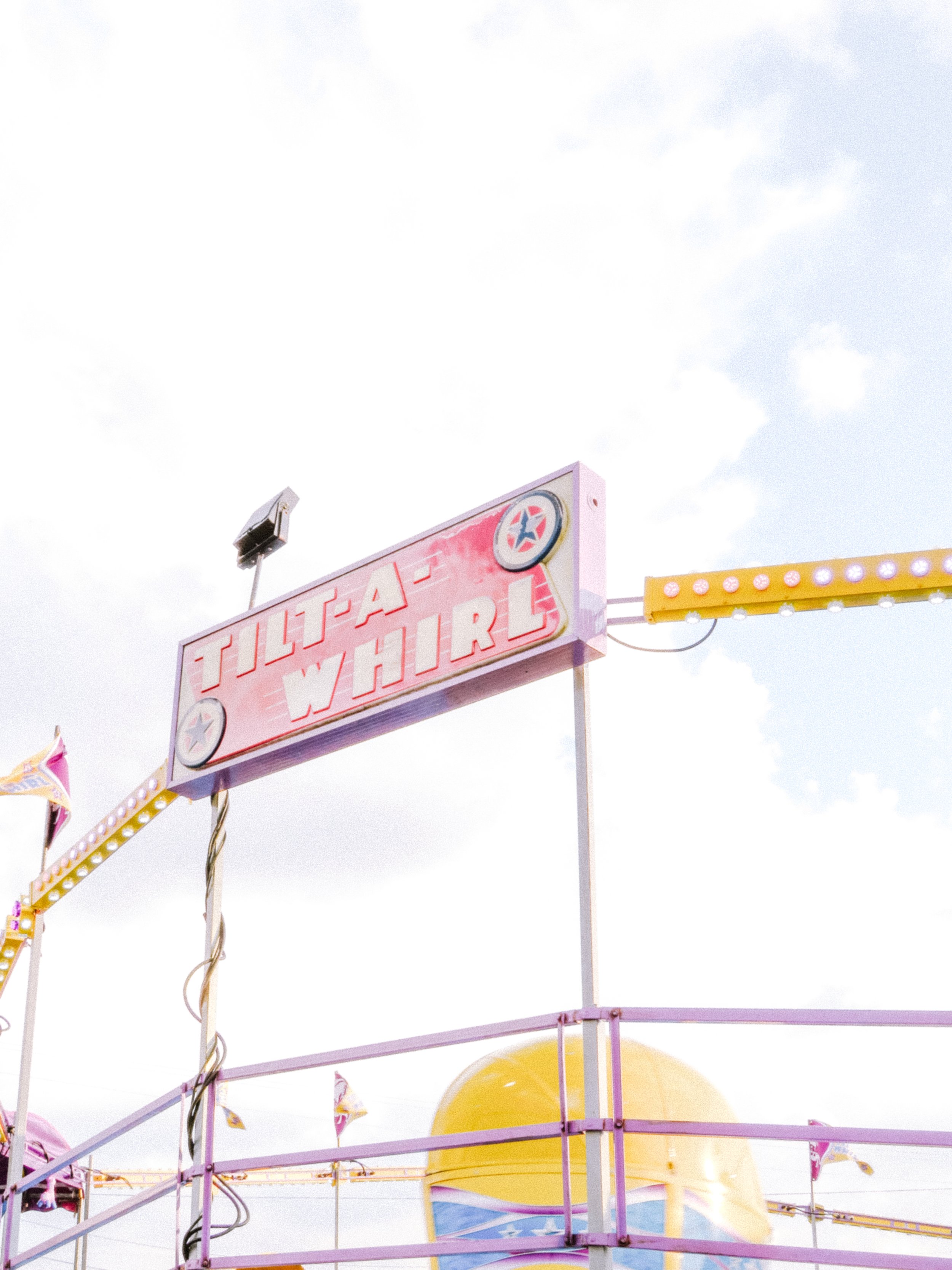 Bright pink sign with the words "FULTA" and "YHI" on a carnival ride. Pink and yellow railings and a large yellow slide are visible, with a cloudy sky in the background.